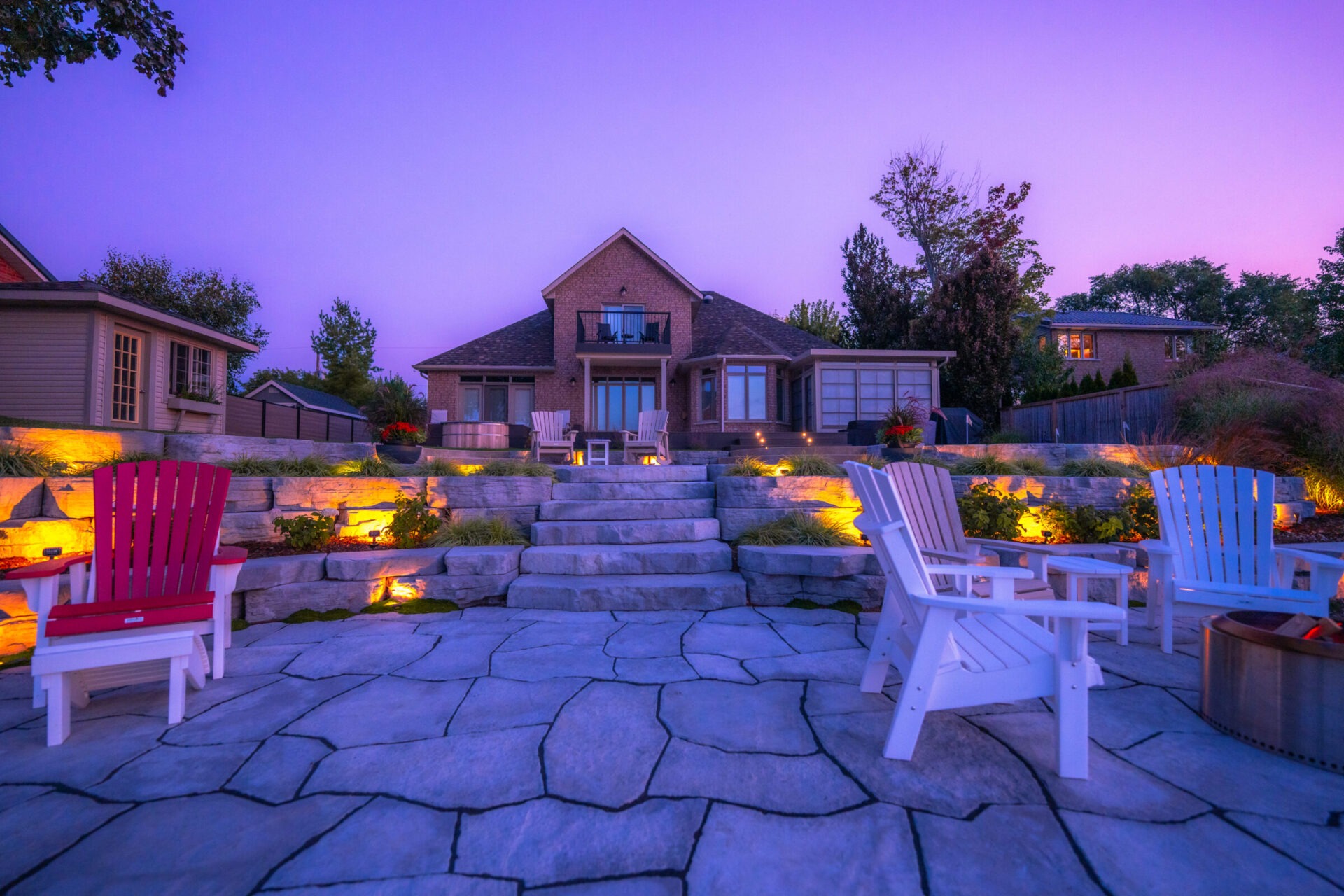 A twilight scene of a landscaped backyard with Adirondack chairs, a fire pit, stone steps, illuminated plant beds, and a cozy brick house.