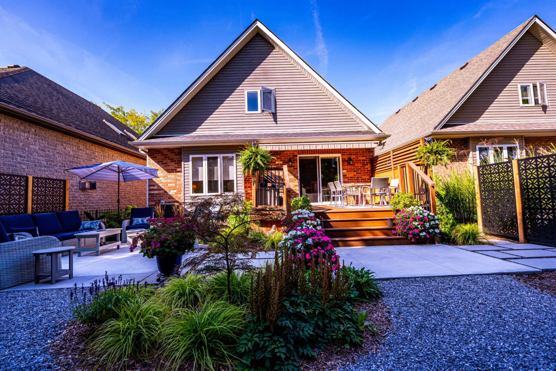 This image shows a cozy suburban home with a wooden deck, outdoor furniture, blooming flowers, and a pebble pathway under a clear blue sky.