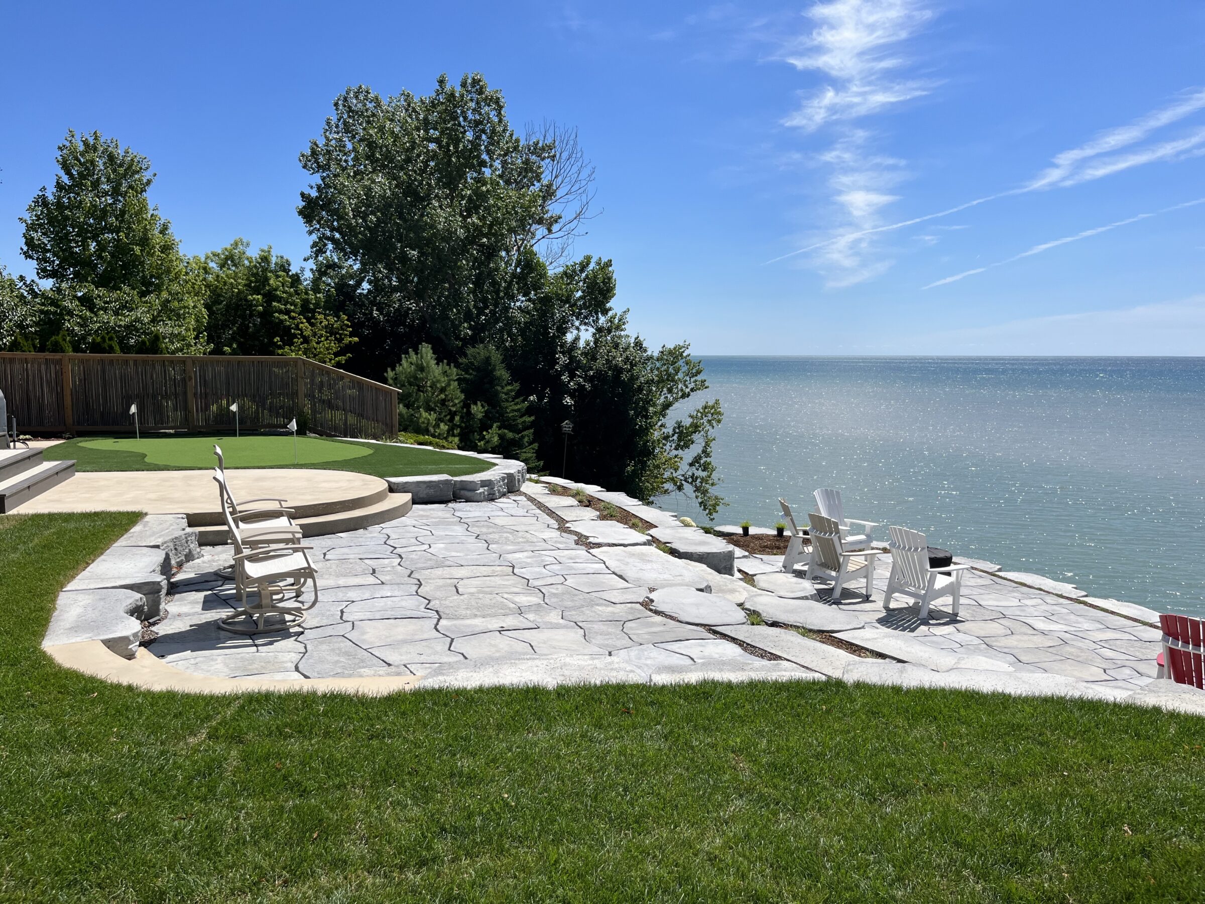 A serene backyard with stone patio, Adirondack chairs overlooking a calm lake, lush green grass, clear blue sky, and a fenced-off putting green area.