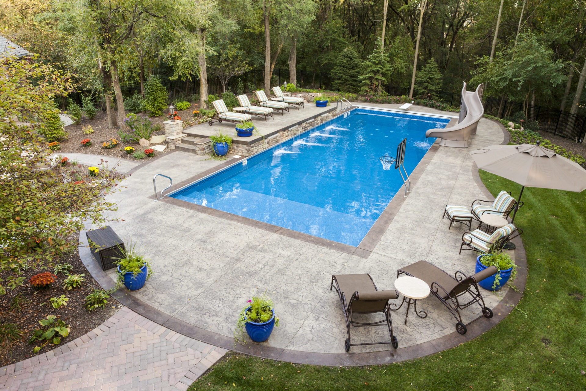 An outdoor residential area featuring a large rectangular pool, lounge chairs, a waterslide, umbrellas, patio furniture, and surrounding green trees.