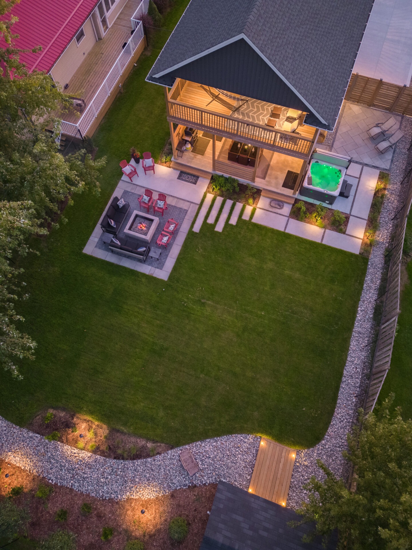 Aerial view of a backyard with a well-maintained lawn, elegant patio furniture, outdoor fireplace, pathway, and a hot tub, at dusk.