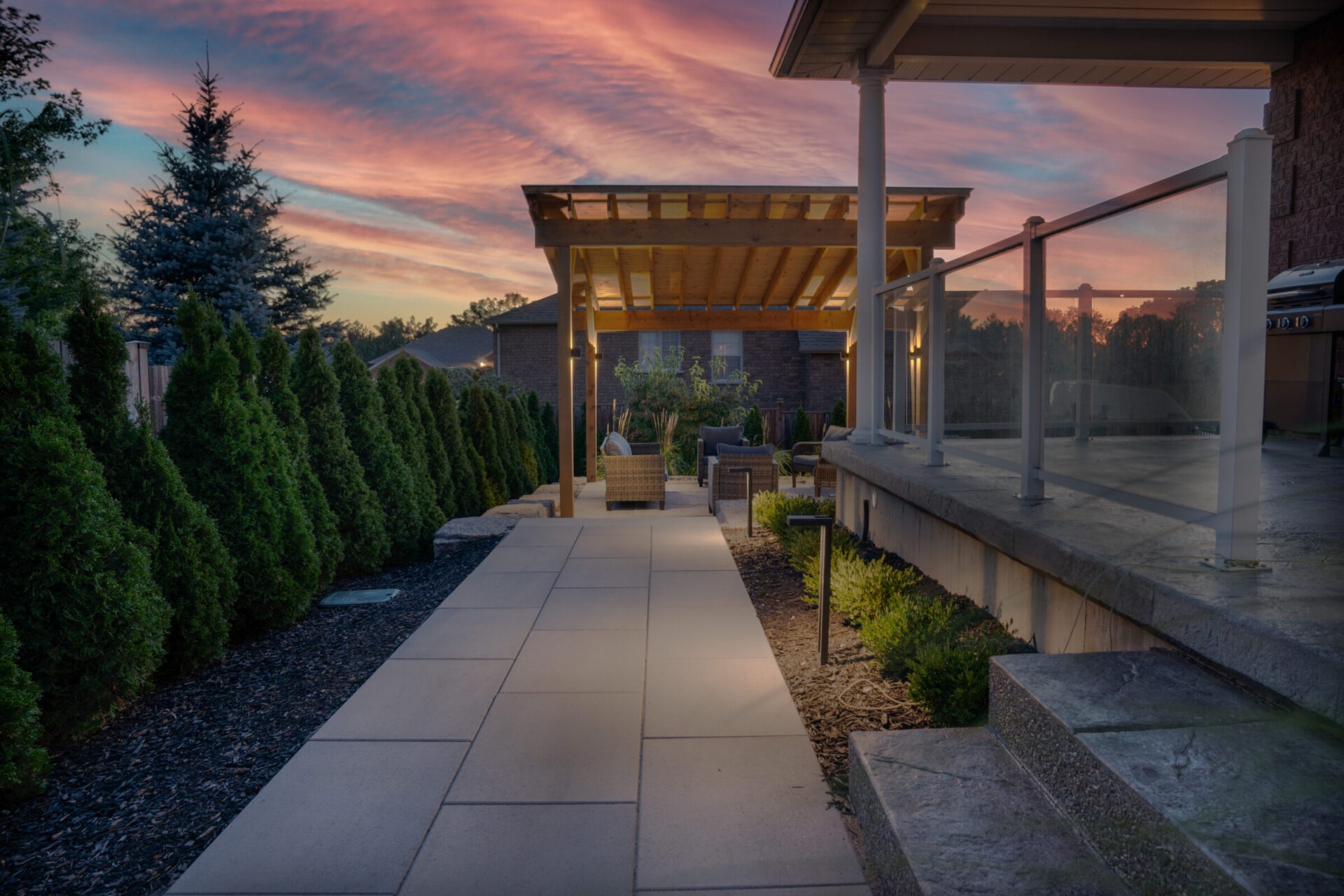 A serene residential patio at dusk featuring outdoor furniture under a wooden pergola, flanked by green shrubbery with a vibrant sunset sky.