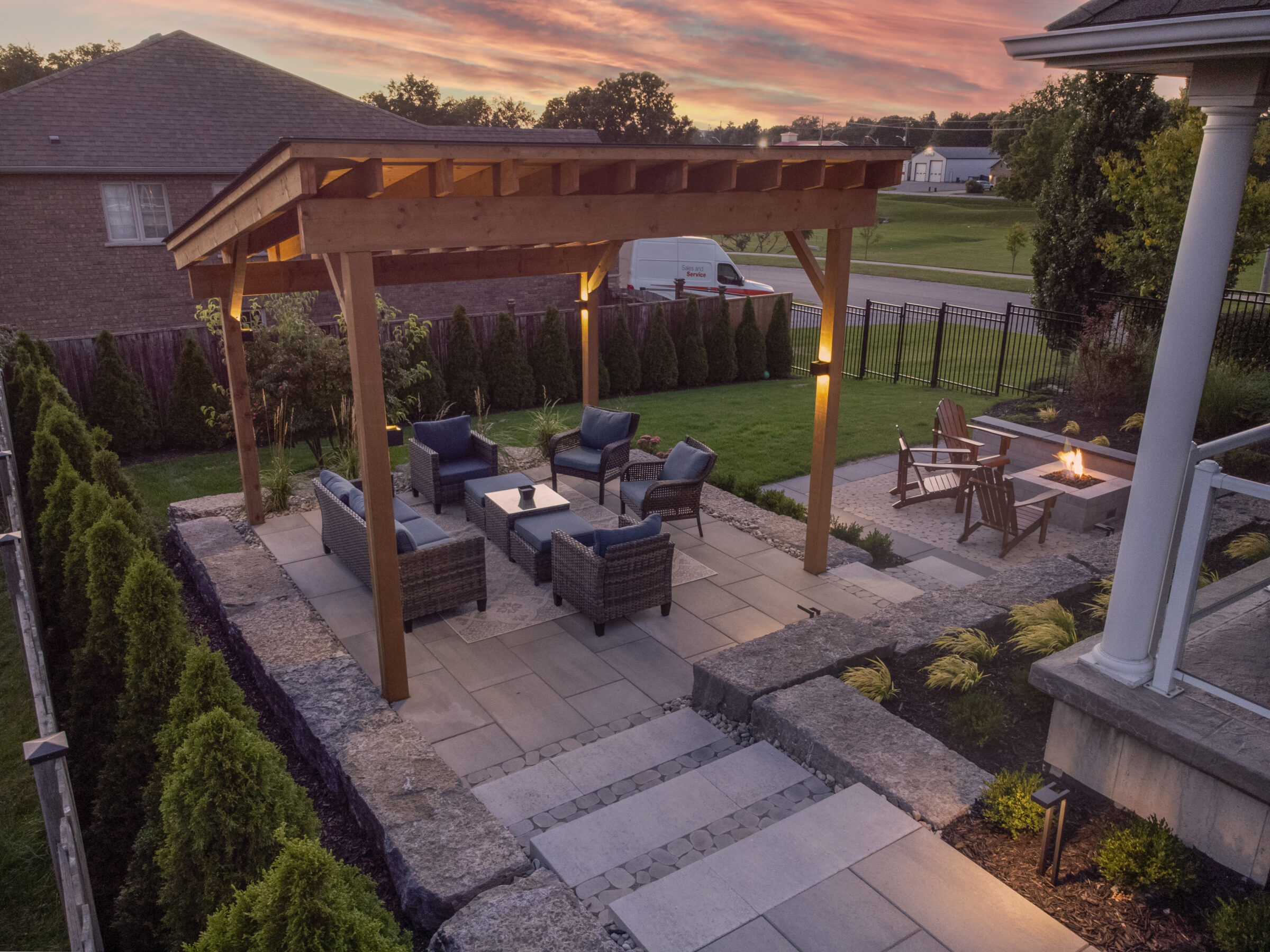 An elevated view of a backyard at dusk, featuring a pergola, outdoor furniture, a fire pit, neatly manicured landscaping, and ambient lighting.