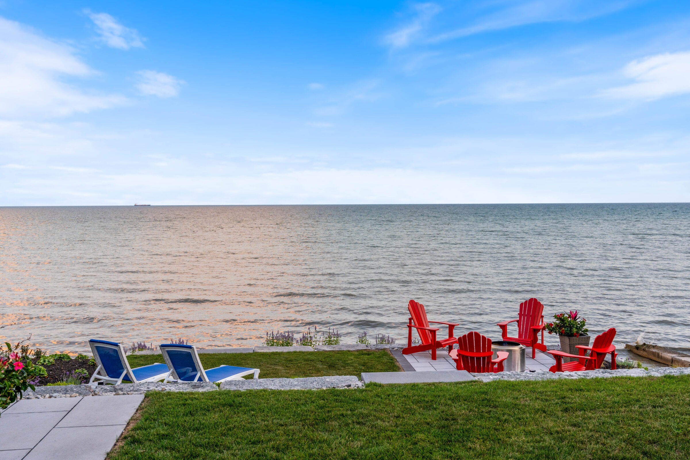 A serene waterfront view with four red Adirondack chairs, two blue loungers, green grass, calm blue water, clear sky, and a distant ship.