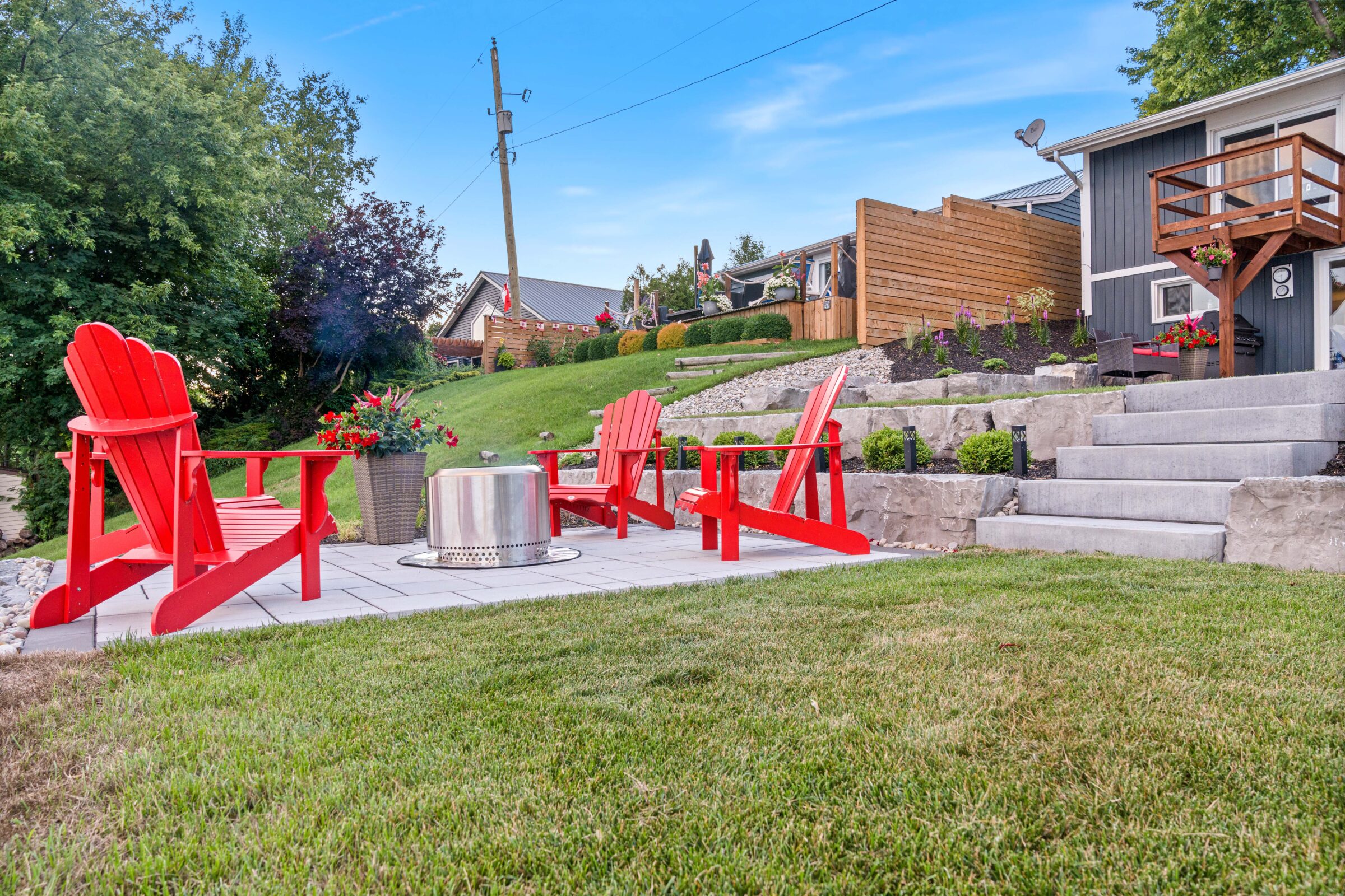 A backyard space with red Adirondack chairs, a fire pit, stone steps, and landscaped grass under a clear blue sky. Residential area with houses.