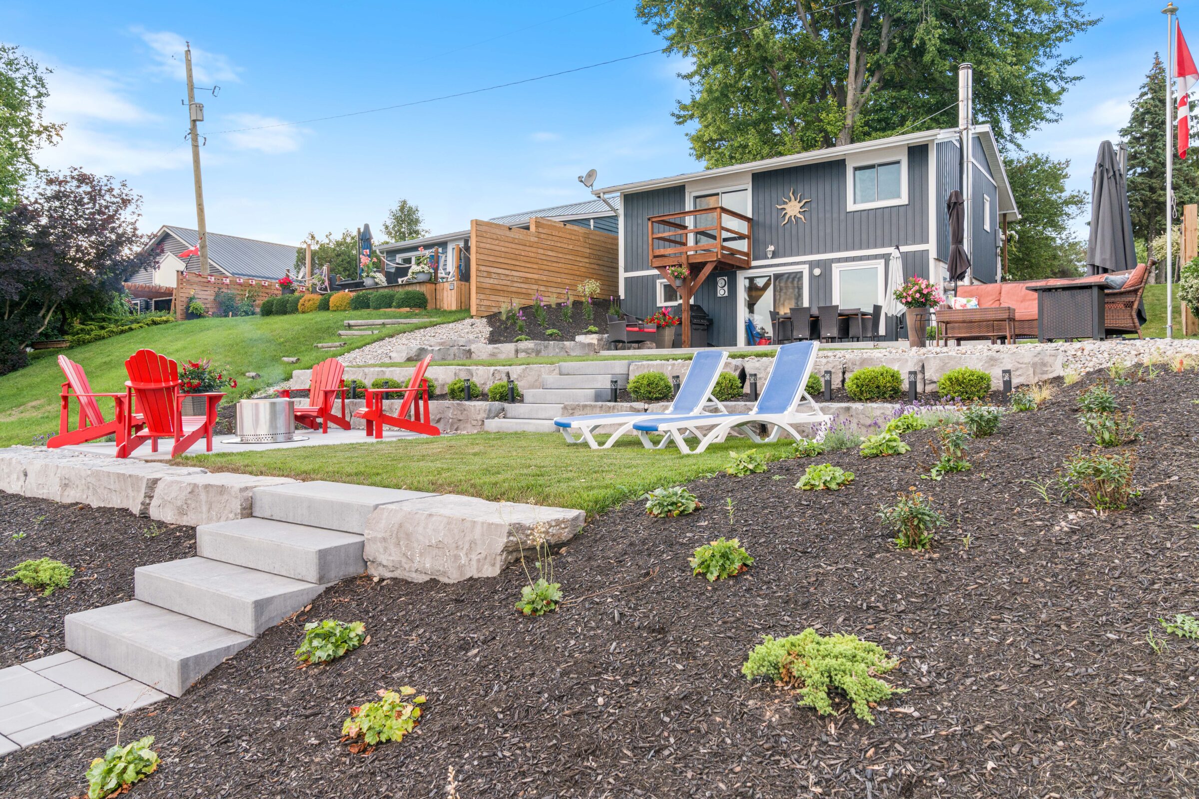A landscaped backyard with red chairs, sun loungers, a fire pit, and a two-story house with a deck. A flag pole and green lawn are visible.