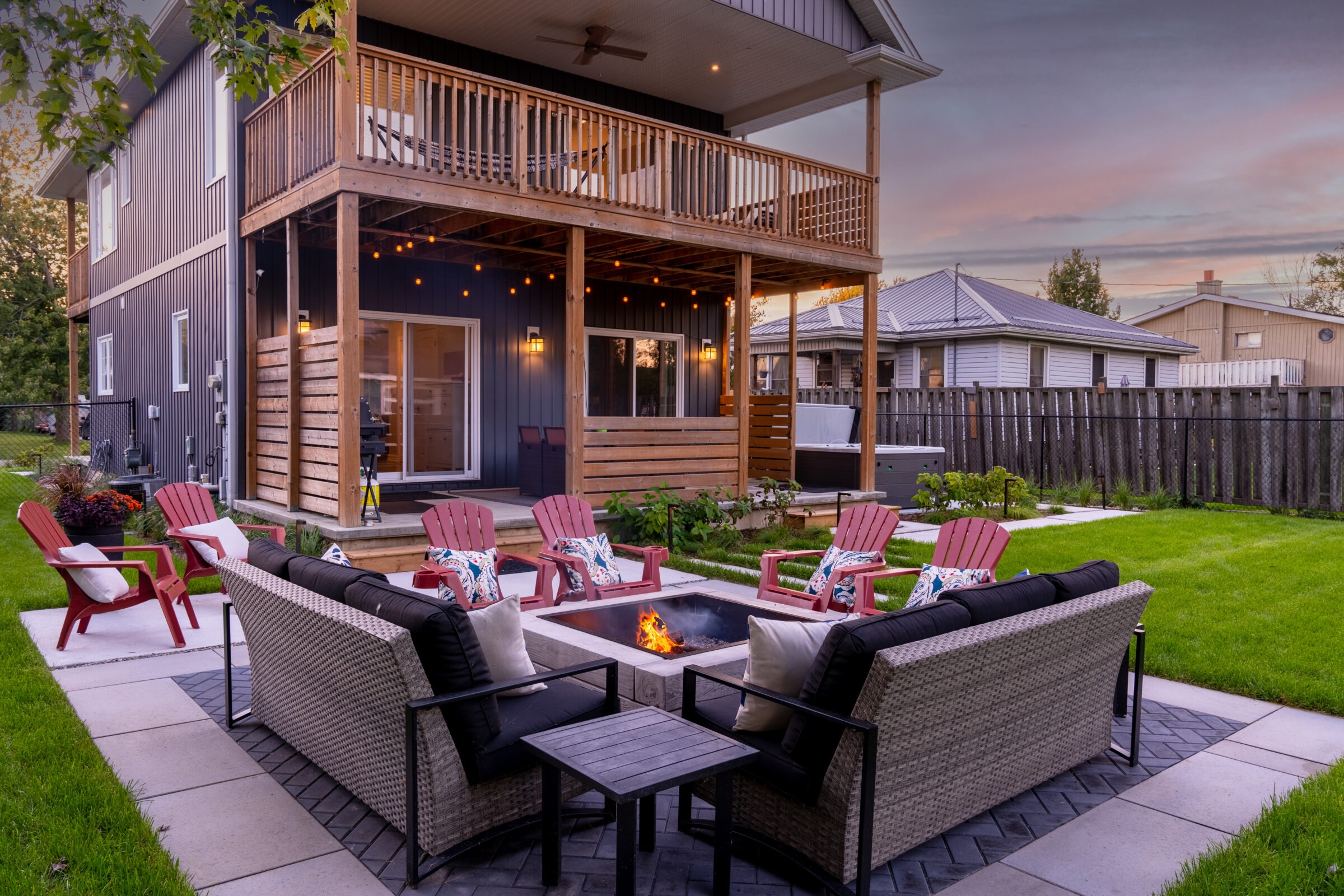 A cozy, well-landscaped backyard with a fire pit, seating area, and a two-story house with wooden accents during dusk. Decorative lights add a warm ambiance.