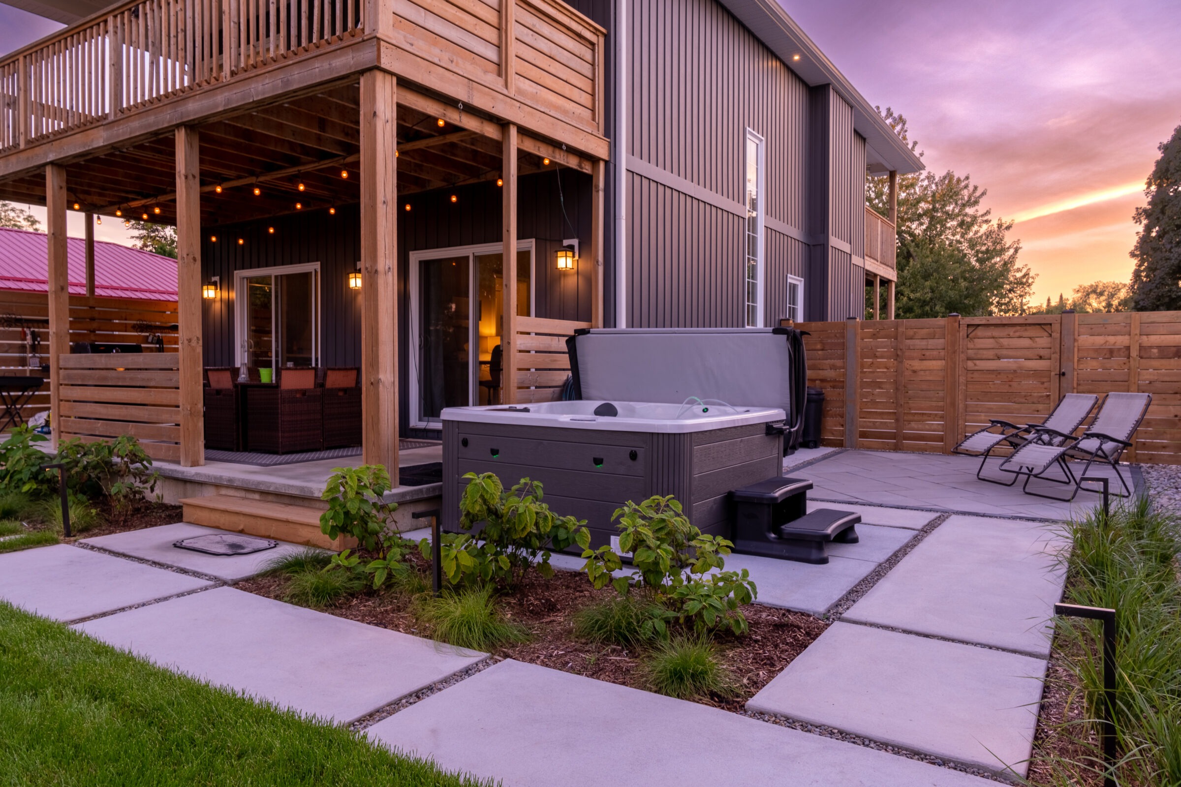 A modern backyard at dusk with a hot tub, lounge chairs, ambient lighting, wooden deck, and landscaping, enclosed by a wooden fence.