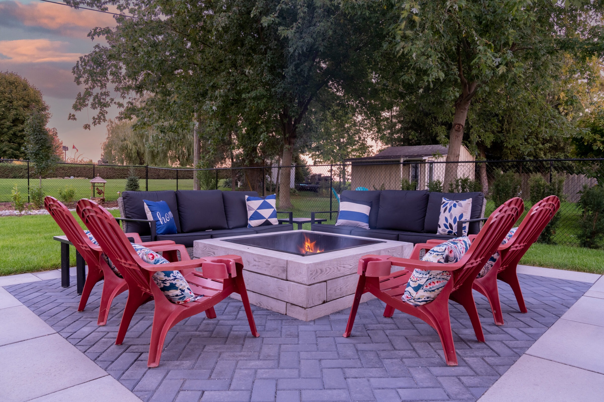 An outdoor patio at dusk with a lit fire pit, surrounded by red chairs and a sofa with decorative pillows, overlooking a green yard.