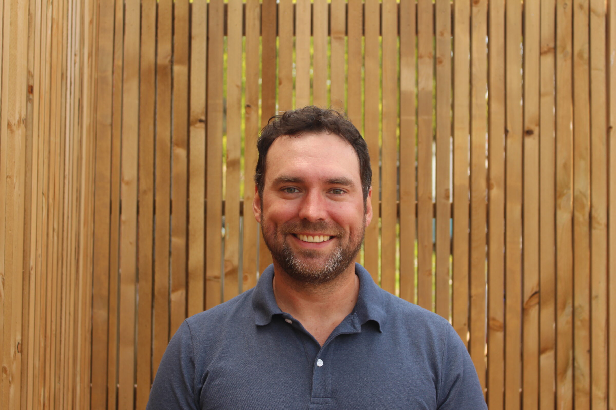 A smiling person stands before a wooden slat background with sunshine filtering through, wearing a polo shirt, exuding a friendly and approachable demeanor.