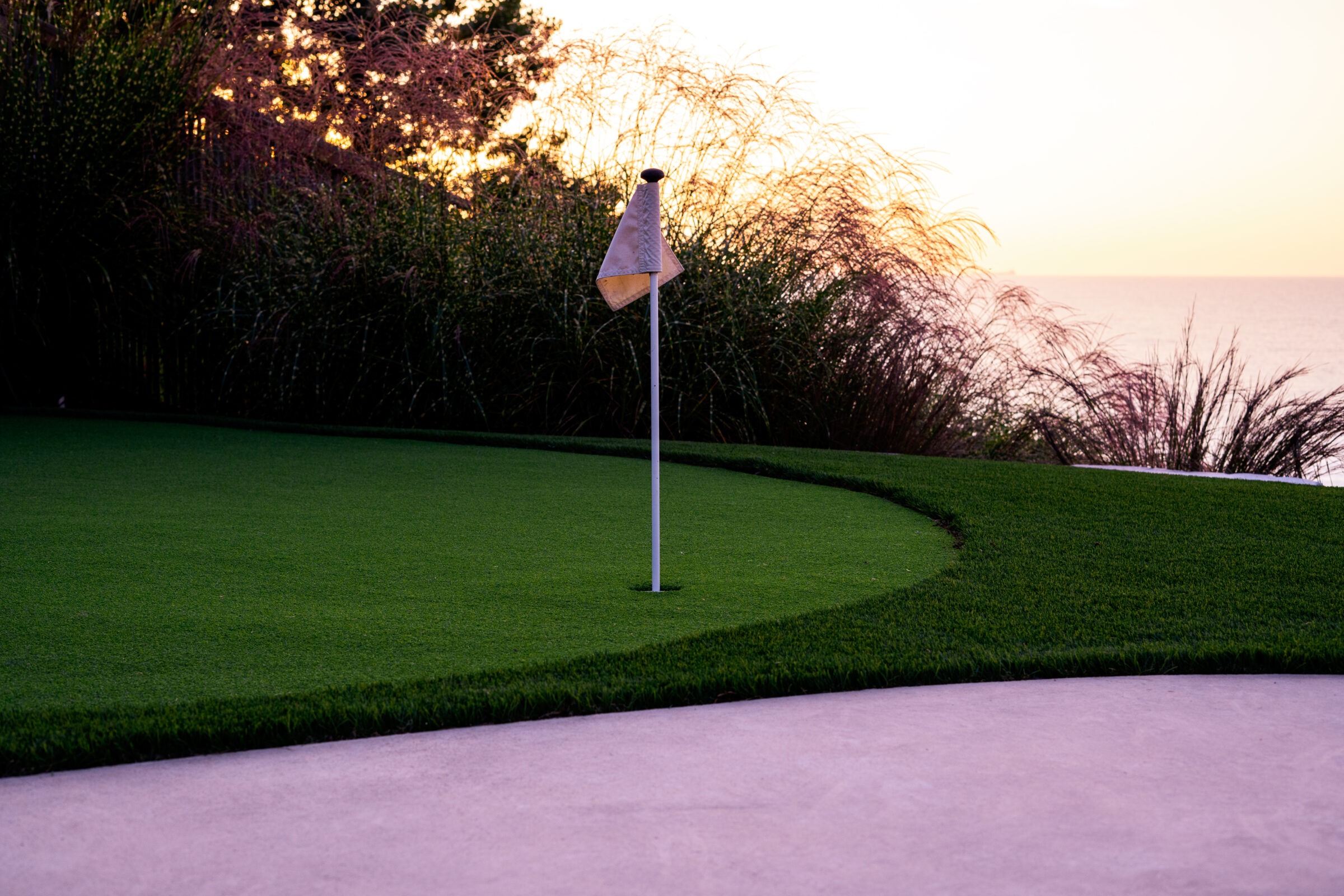 A golf hole with a flagstick on a green, bordered by tall grasses, with a sunset and the ocean horizon in the background.