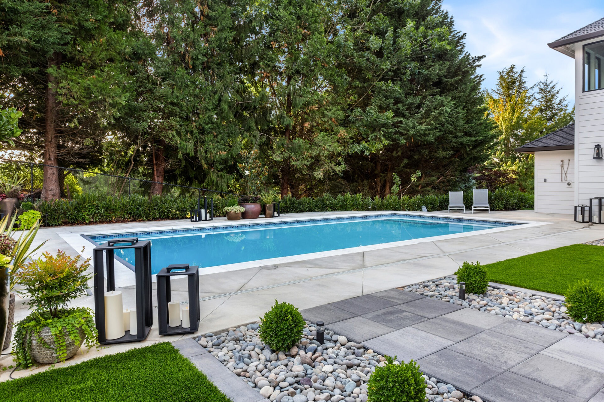 A tranquil backyard with a rectangular swimming pool, neat landscaping, lounge chairs, and tall trees under a clear sky during the day.