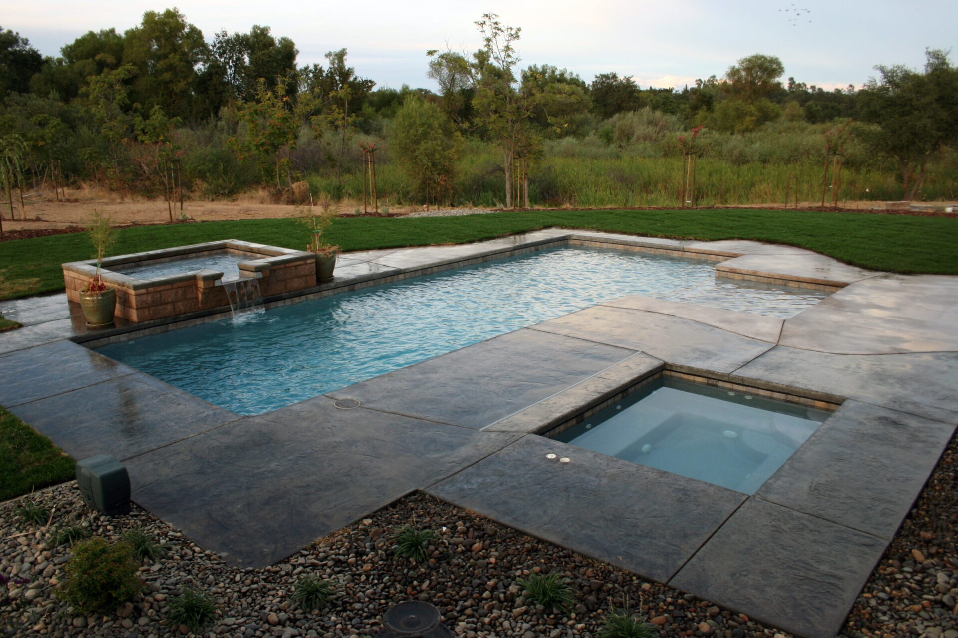 This image shows a luxurious backyard with a rectangular swimming pool, an attached raised spa with flowing water, and lush greenery in the background.