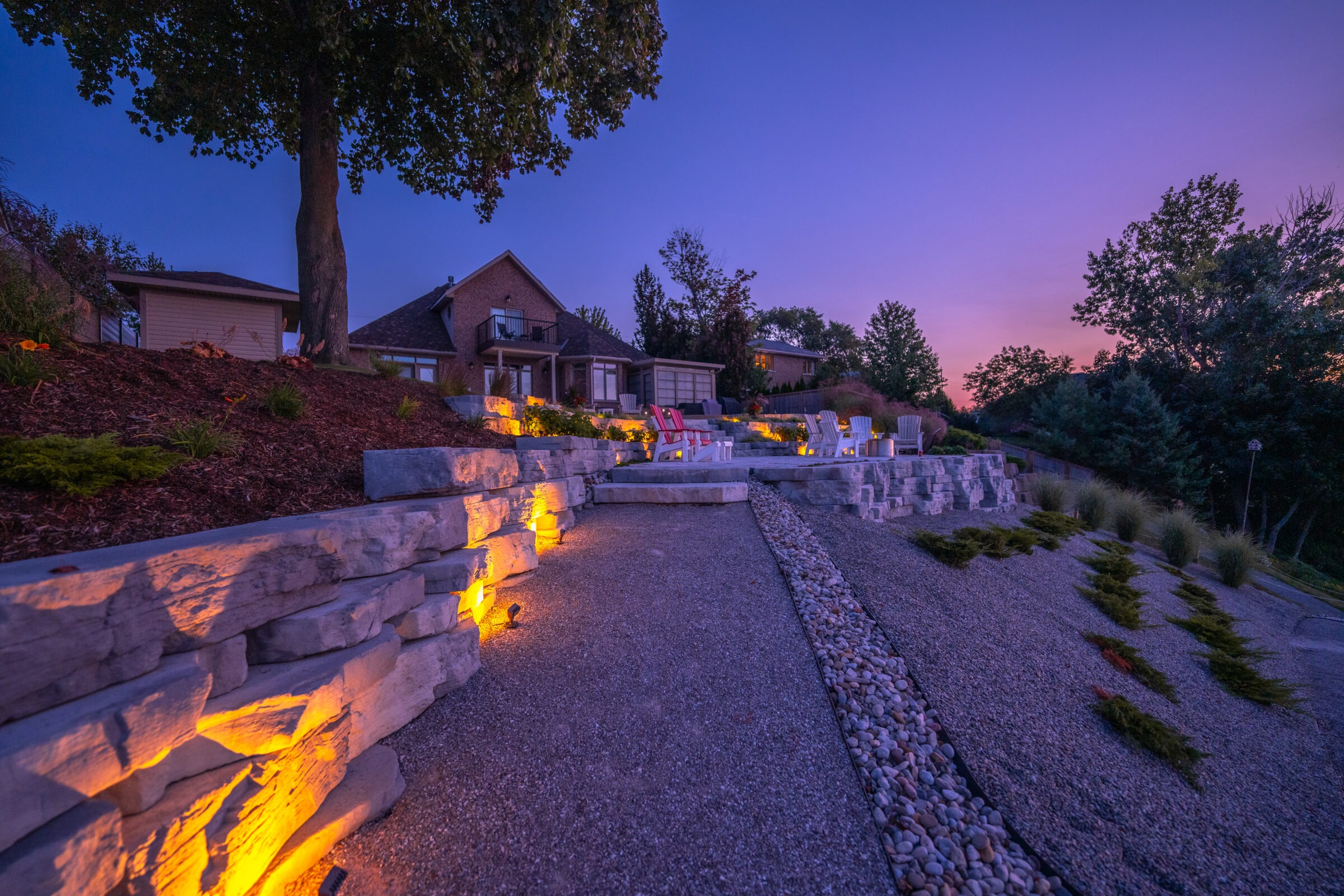 A serene dusk setting featuring a landscaped backyard with lit stone retaining walls, a gravel path leading to Adirondack chairs, and a tranquil sky.