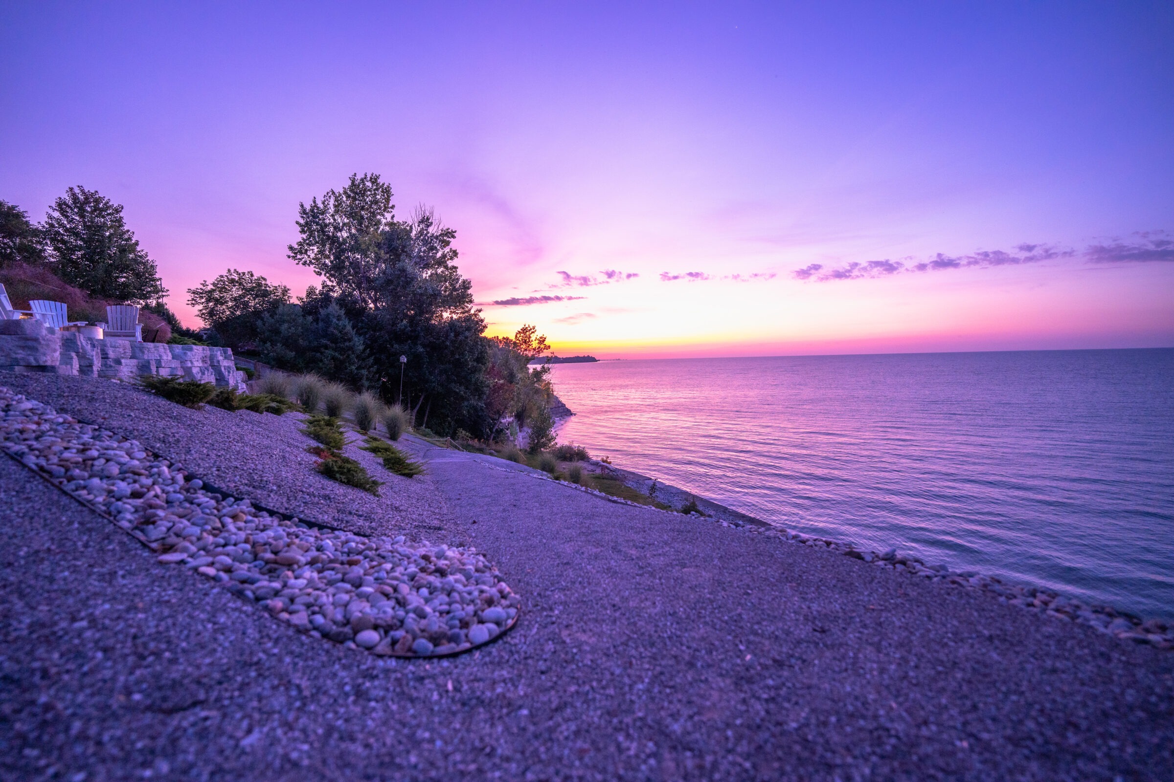A tranquil lakeside at twilight with Adirondack chairs overlooking calm waters, accentuated by a sky with hues of purple and orange from the setting sun.