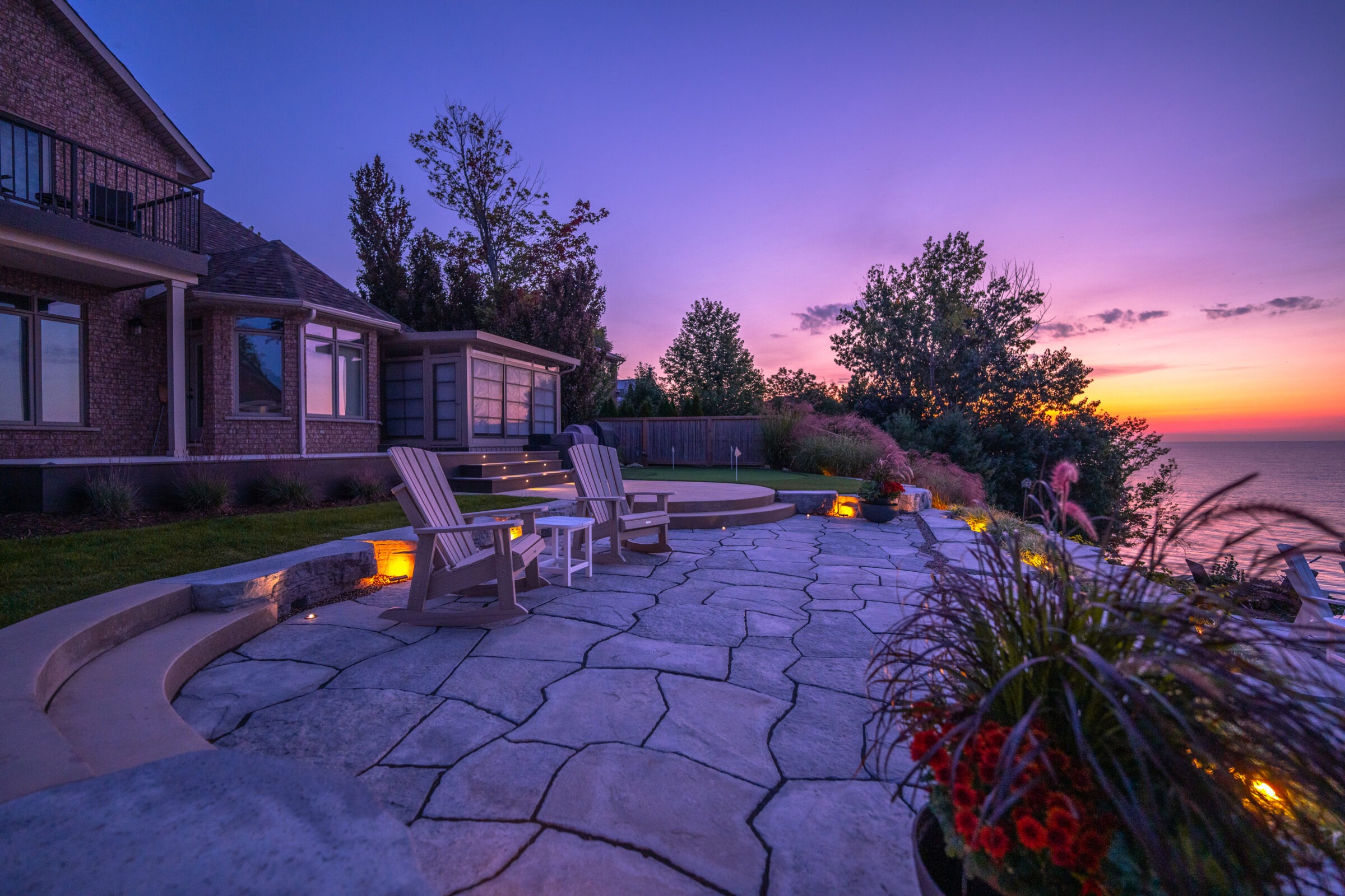 A tranquil backyard with an ocean view at sunset features Adirondack chairs, elegant landscaping, a fire pit, and a stone patio adjoining a brick house.