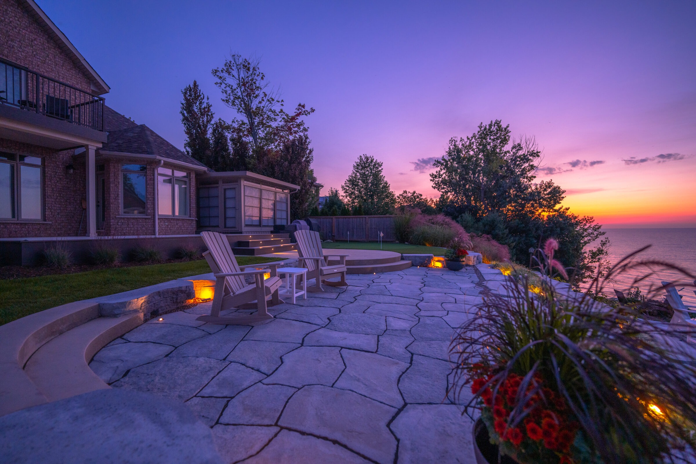 A serene dusk setting with a purple sky at a luxurious backyard patio overlooking the sea, featuring Adirondack chairs and elegant landscaping.