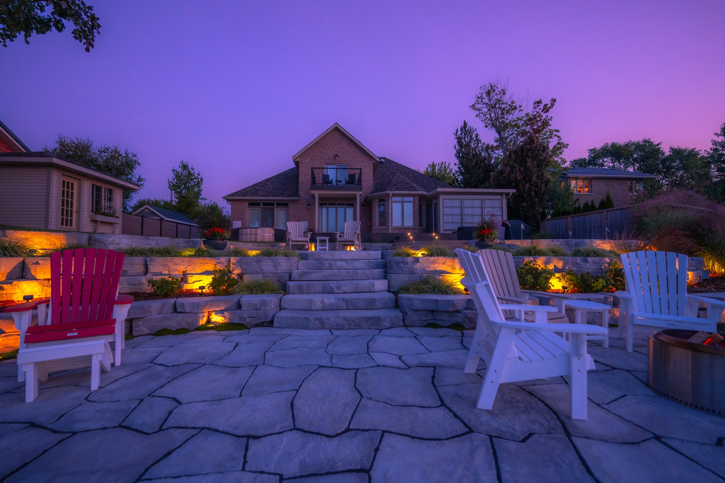 An elegant backyard during twilight with lighting accentuating a stone patio, Adirondack chairs, landscaping, and a brick house under a purple sky.
