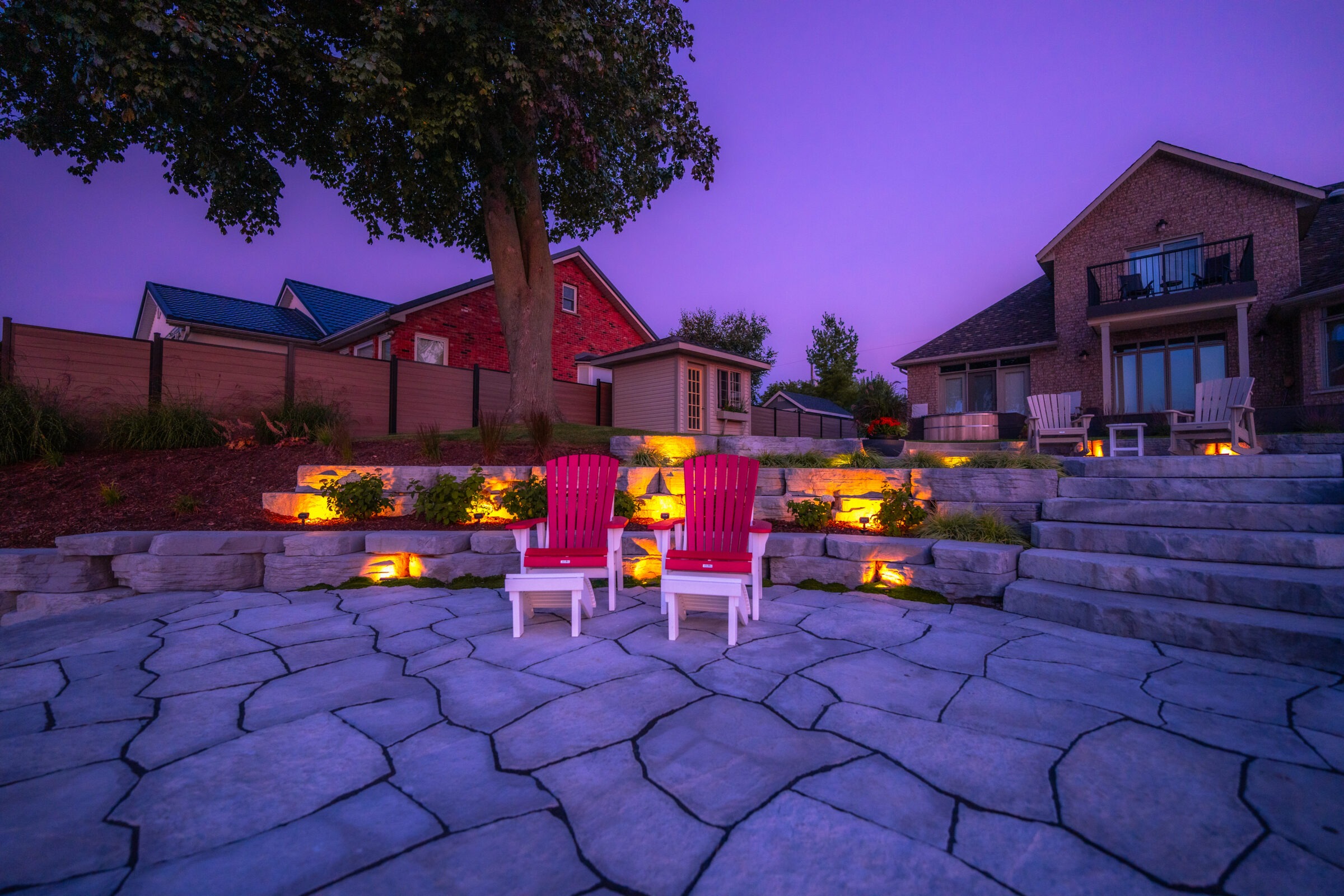 Two red Adirondack chairs on a stone patio with integrated lighting, stone steps, and a landscaped garden against a twilight sky.