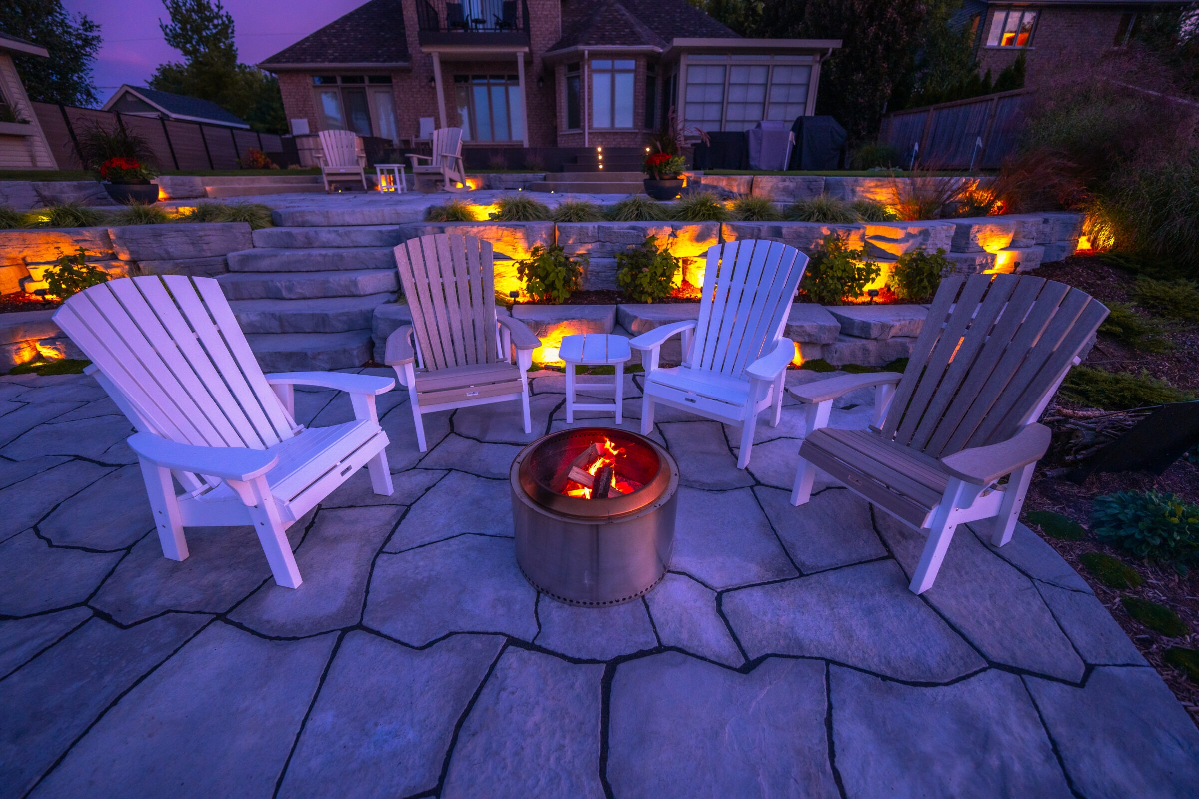 An outdoor scene at dusk with Adirondack chairs surrounding a fire pit, on a stone patio, with illuminated landscaping and a house in the background.
