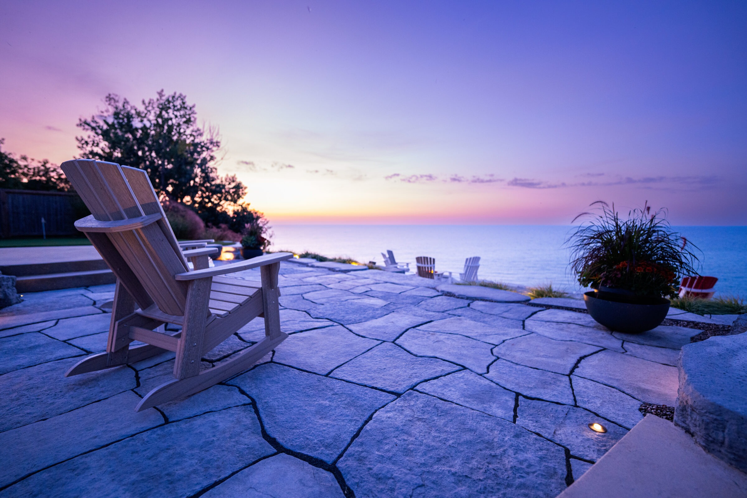 A serene patio overlooks the ocean at sunset with an Adirondack chair in the foreground on stone paving, surrounded by soft evening colors.