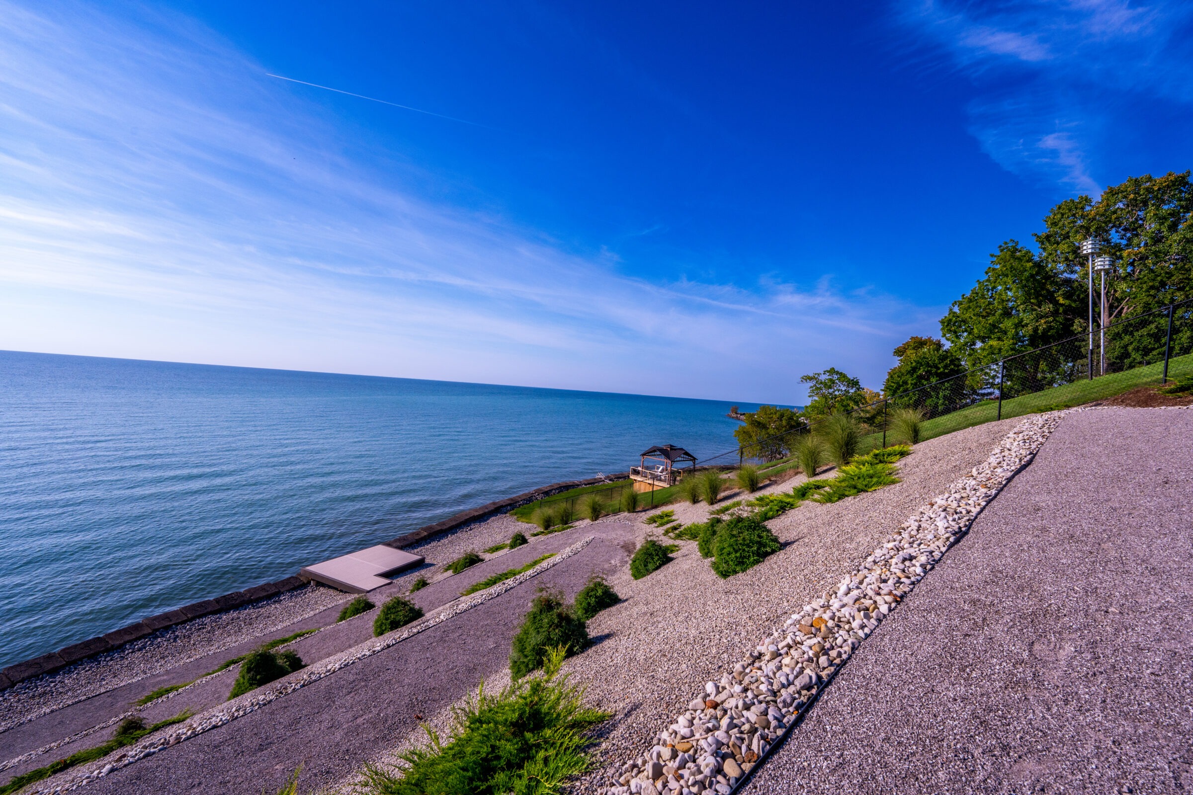 A serene waterfront with tiered landscaping, a gazebo by the shore, clear skies, and a tranquil expanse of water meeting a blue sky at the horizon.