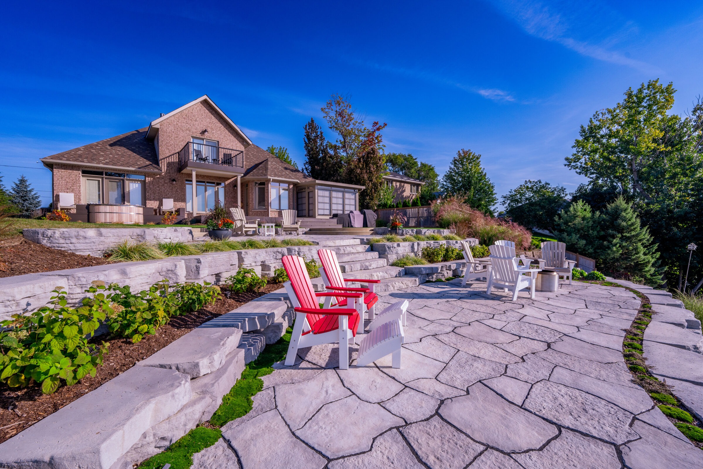 A spacious backyard with a stone patio and tiered garden walls in front of a two-story brick house. Outdoor furniture provides a relaxing seating area.