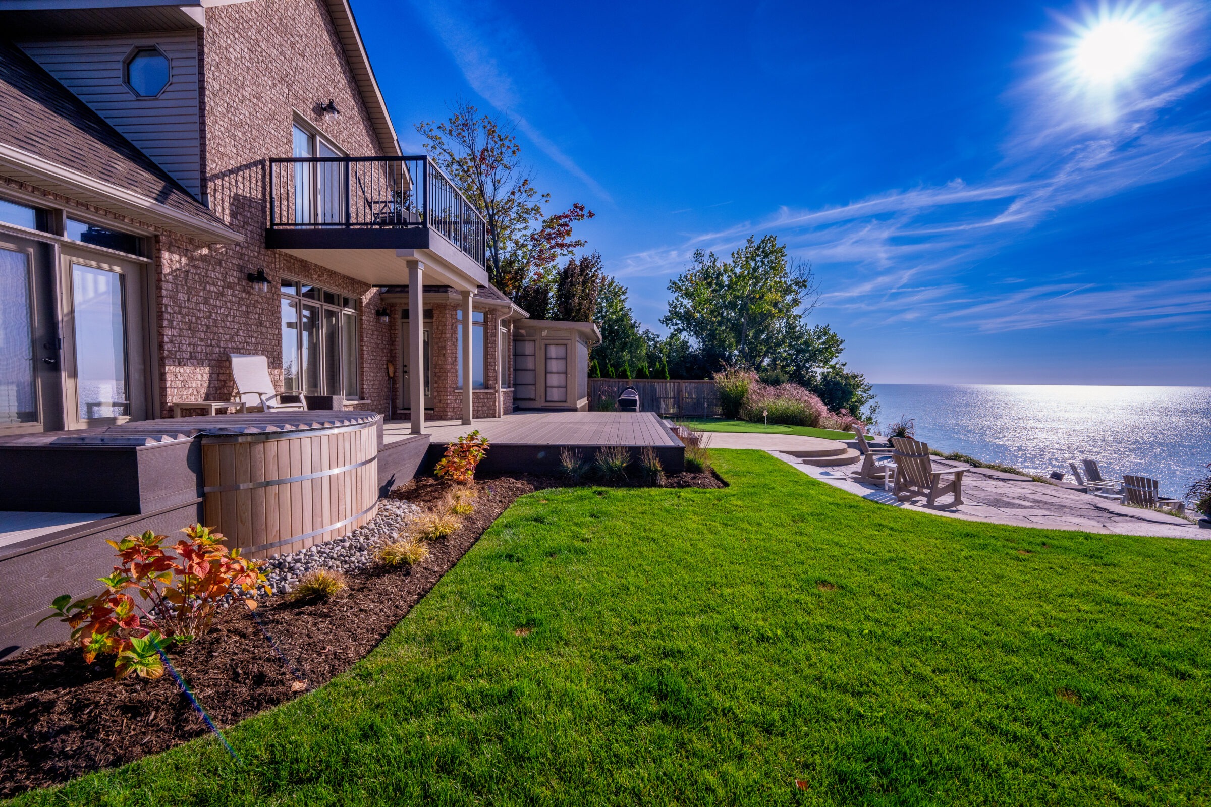 The image shows a luxurious lakeside house with green lawn, outdoor seating, clear skies, a hot tub, and a breathtaking view over the water.
