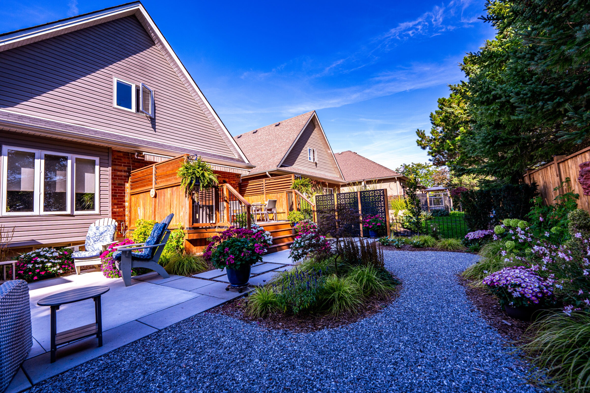 This image shows a beautifully landscaped backyard with a gravel path, colorful flowers, a wooden deck with furniture, and a cozy suburban house.
