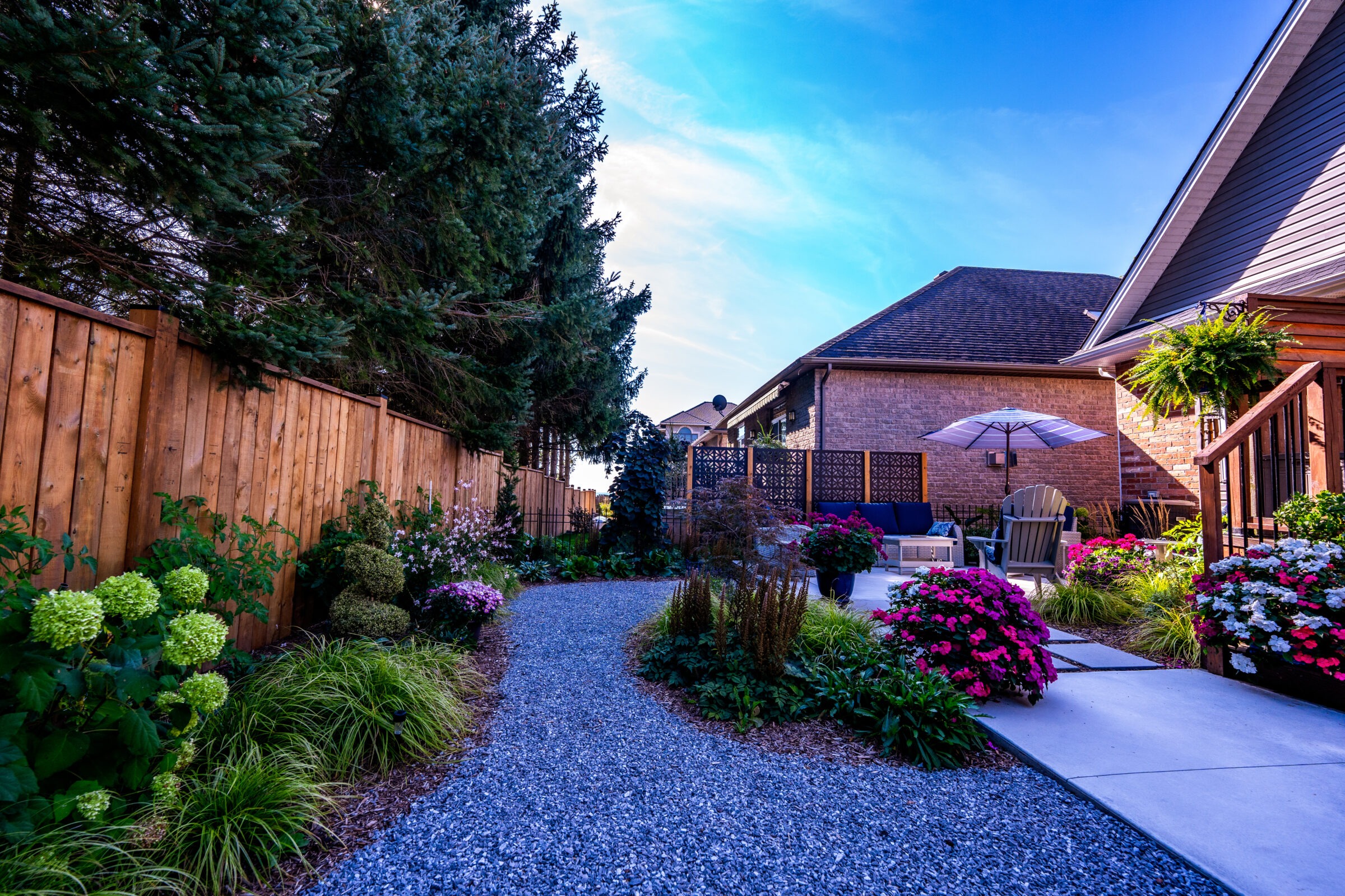 A vibrant garden with a gravel path leading to a cozy patio area adorned with colorful flowers, lush greenery, and a parasol for shade.