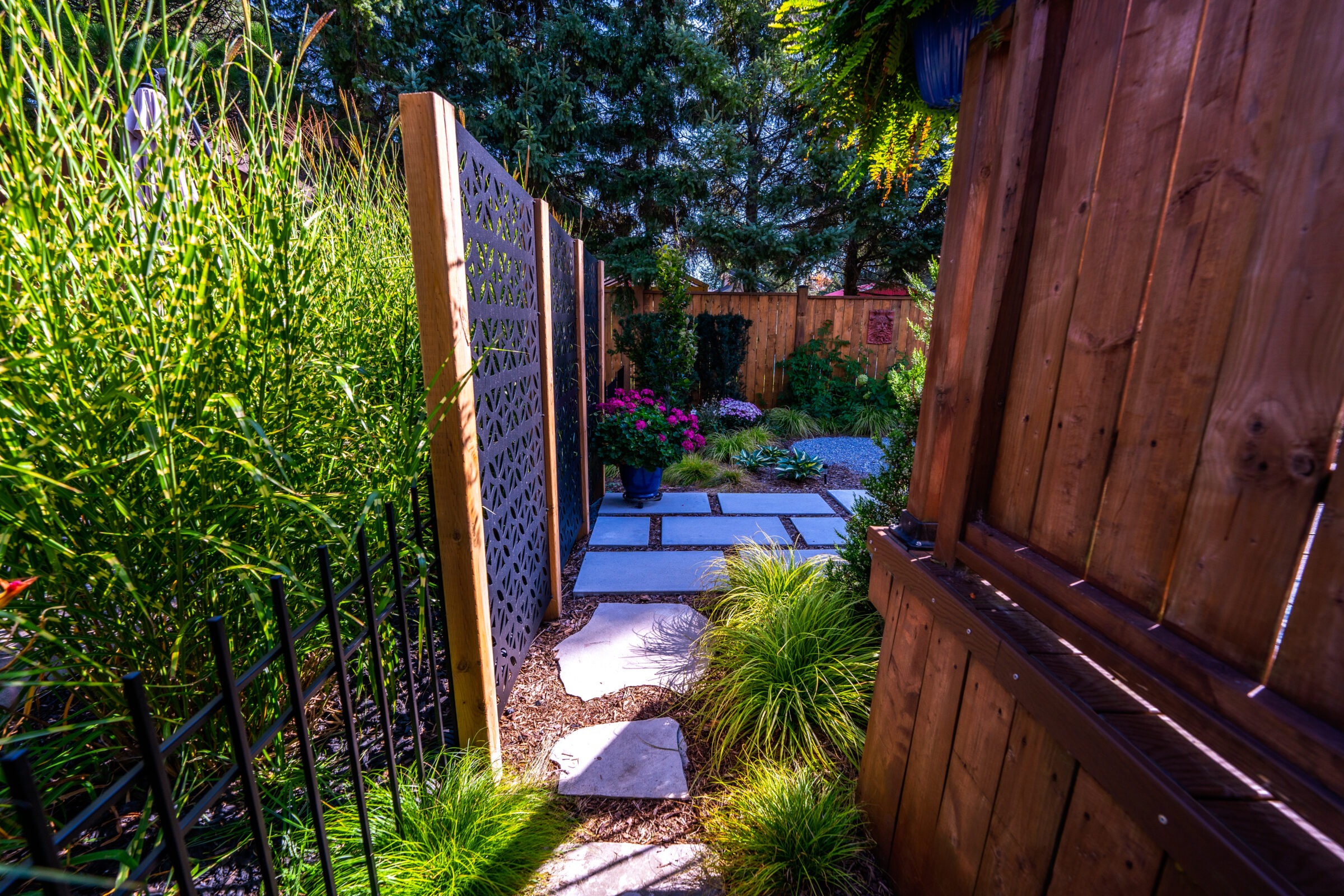 A tranquil garden pathway lined with greenery, stepping stones, a wooden fence, and lush plants. The setting is serene and well-maintained, inviting relaxation.