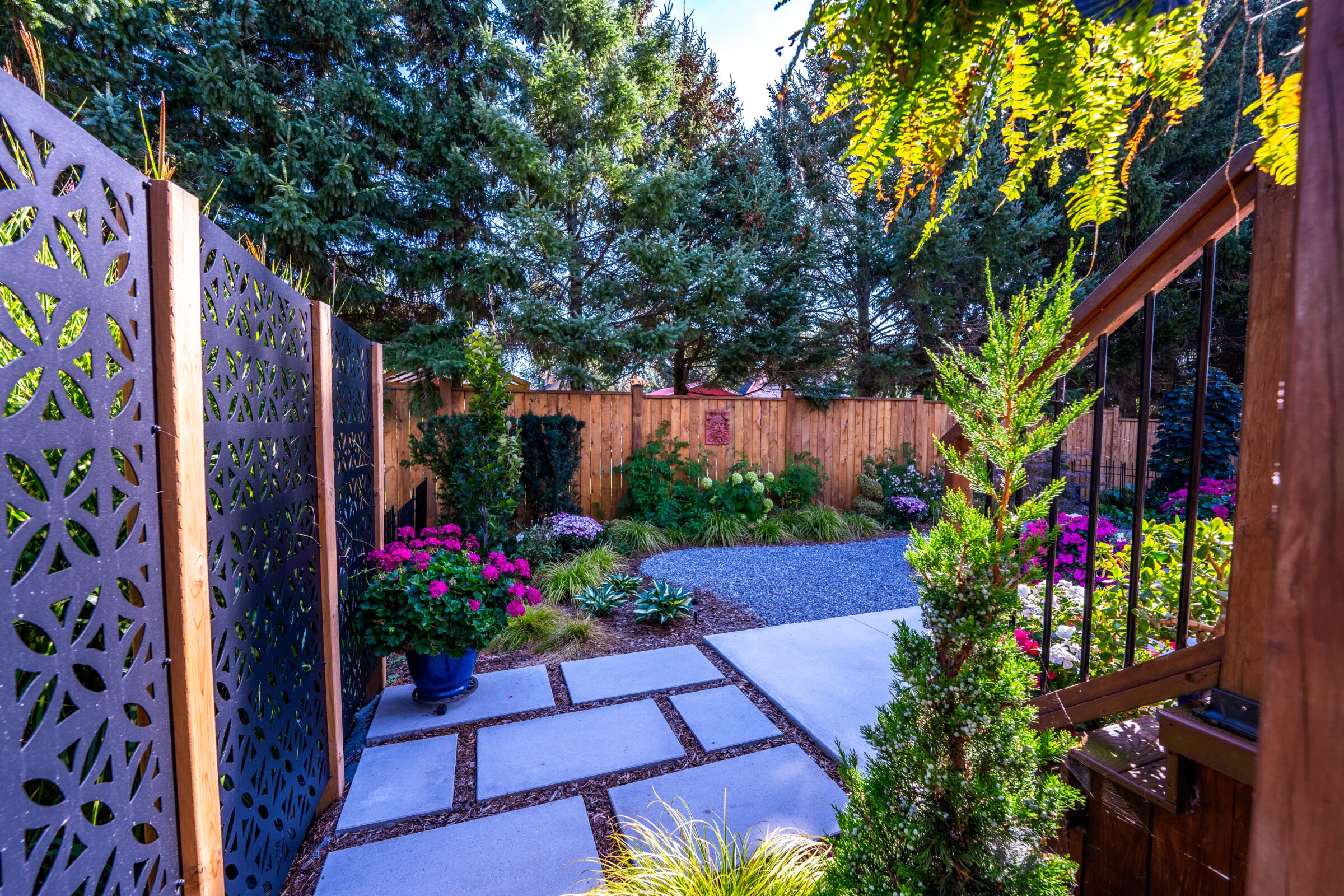 This image shows a well-manicured garden with decorative panels, a wooden fence, gravel path, and an array of colorful flowers and greenery.