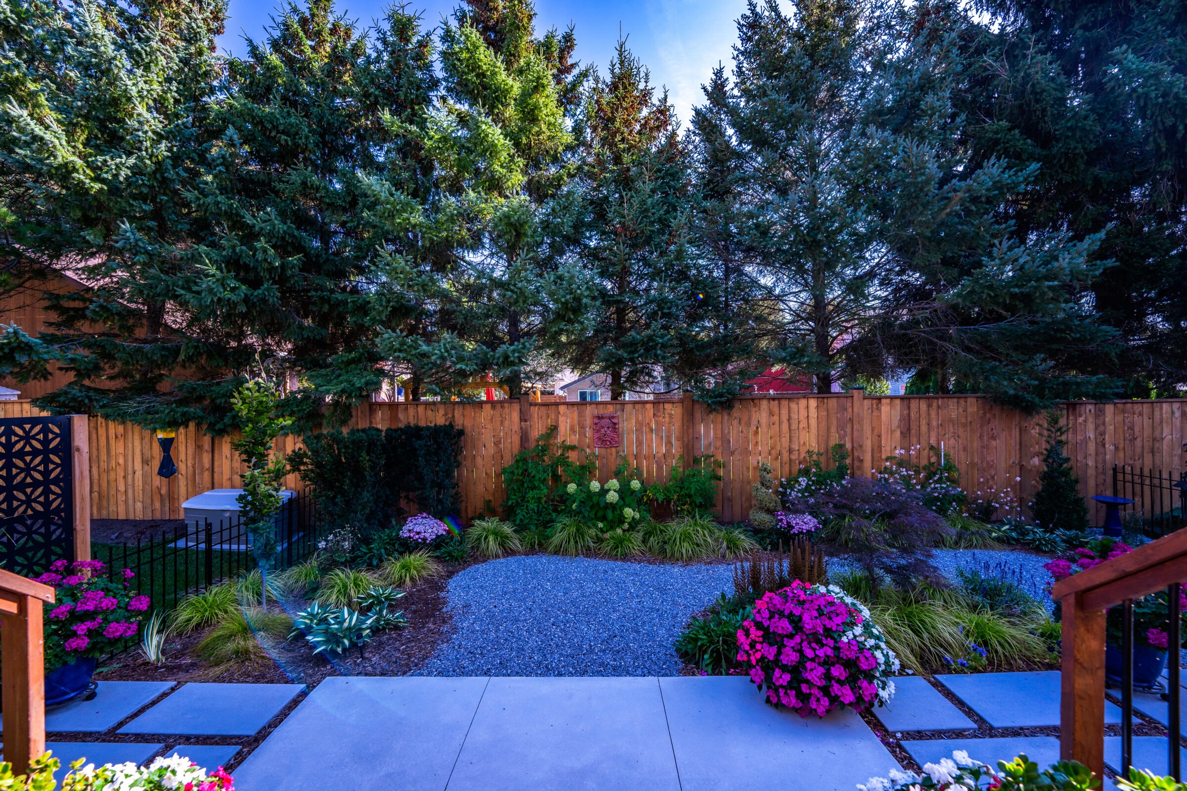 An inviting backyard garden with vibrant flowers, greenery, a gravel path, tall trees, and a wooden fence under a clear blue sky at dusk.