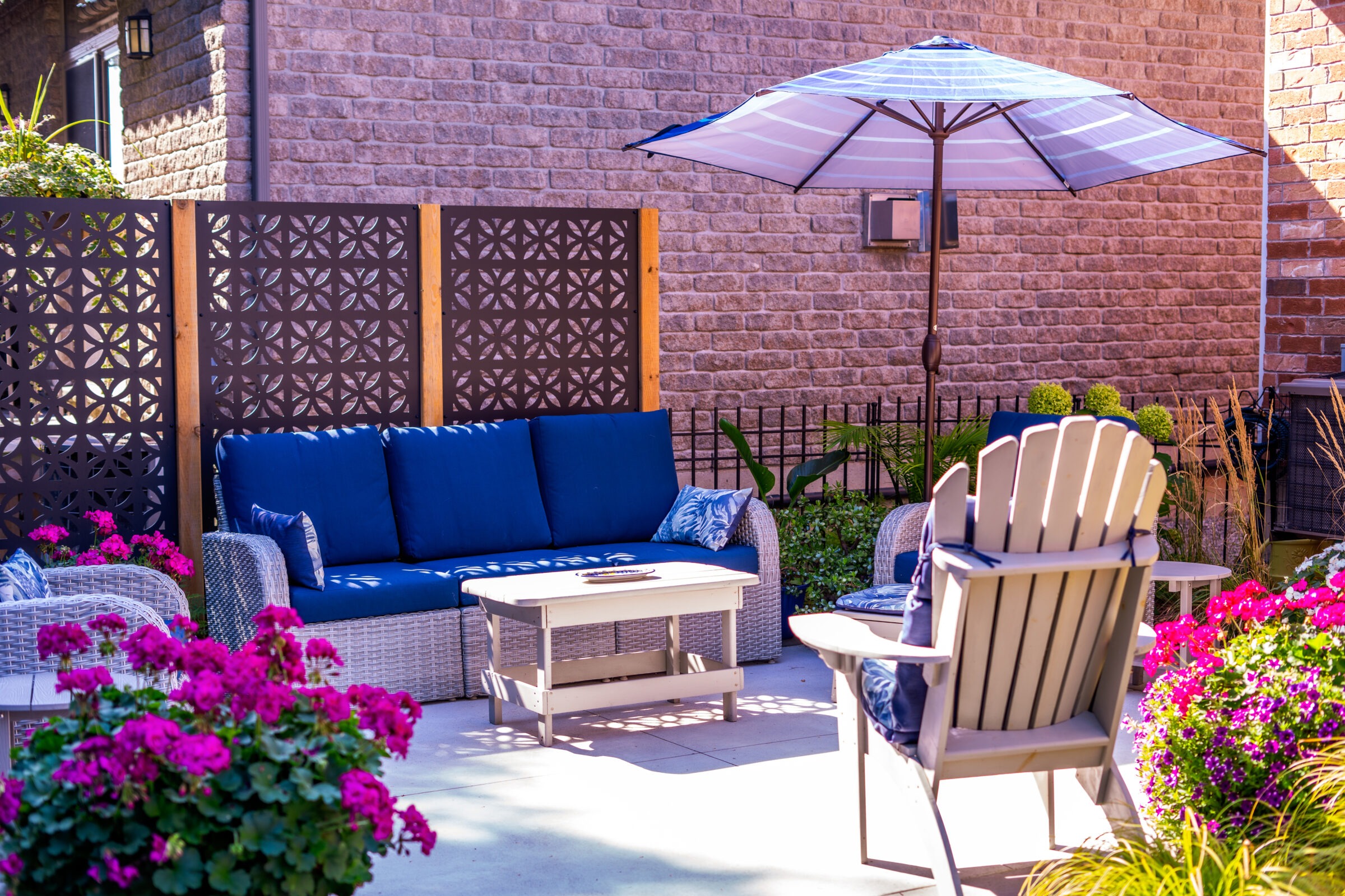 A cozy outdoor patio with a navy cushioned sofa, wicker chairs, and a striped umbrella. Decorative plants and a brick wall enhance the ambiance.