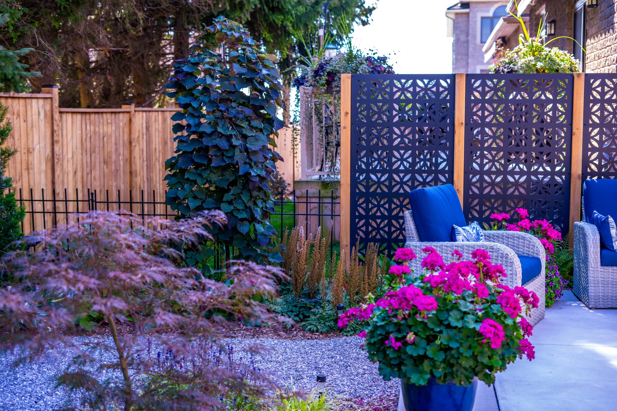The image shows a landscaped garden with a wooden fence, ornate metal panels, lush plants, a pebbled ground, and wicker furniture with blue cushions.