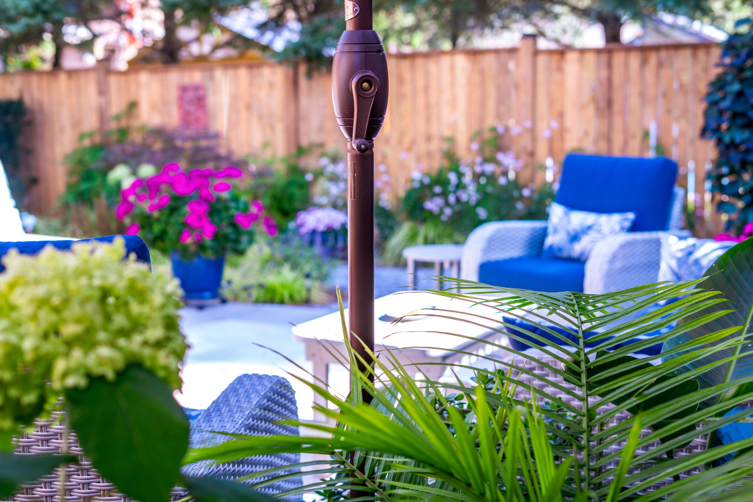 An inviting outdoor patio scene with a close-up of a tiki torch, surrounded by vibrant green plants, comfortable blue-cushioned furniture, and purple flowers.