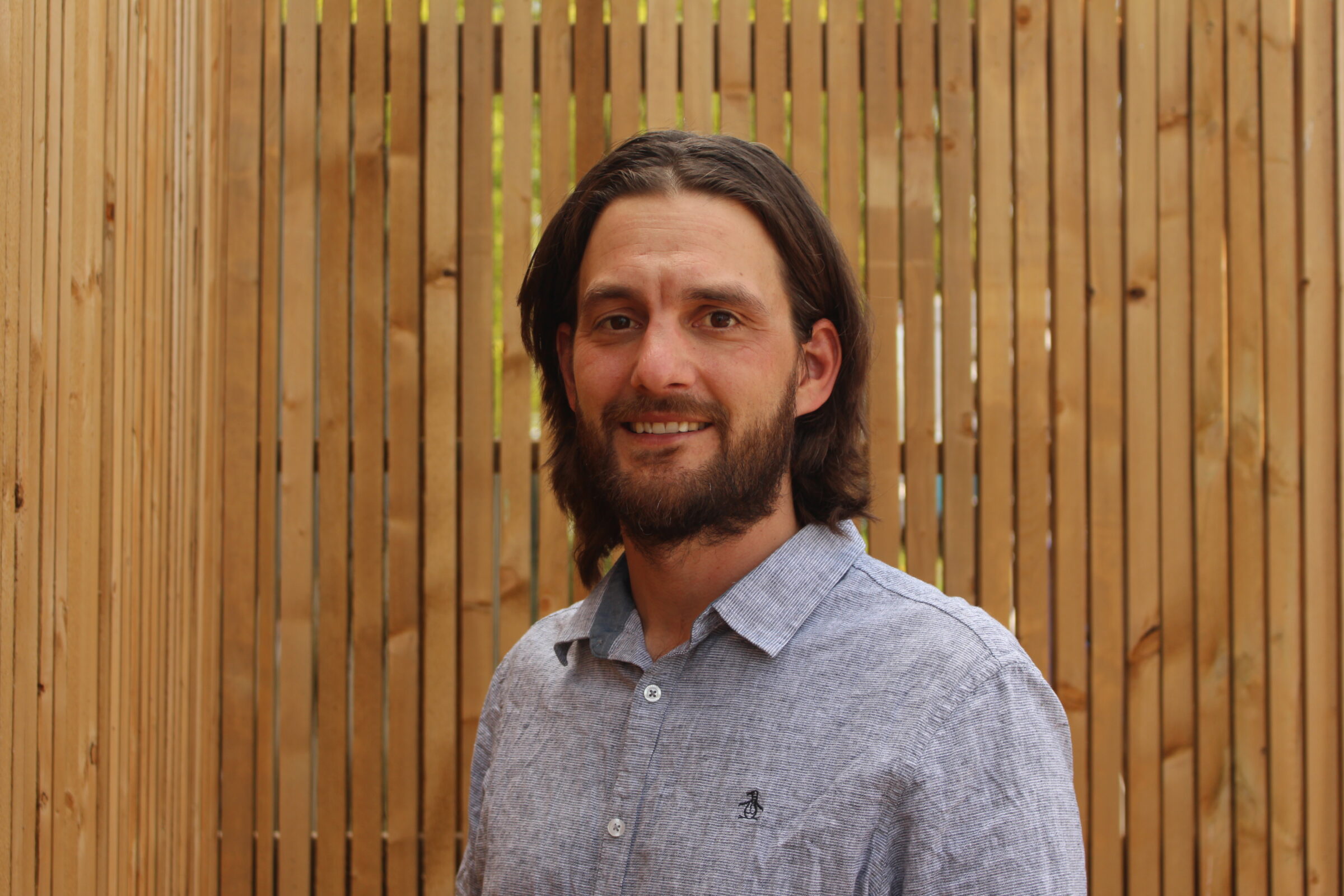 A person is standing in front of a wooden slat background. They are smiling lightly, have medium-length hair, a beard, and are wearing a button-up shirt.