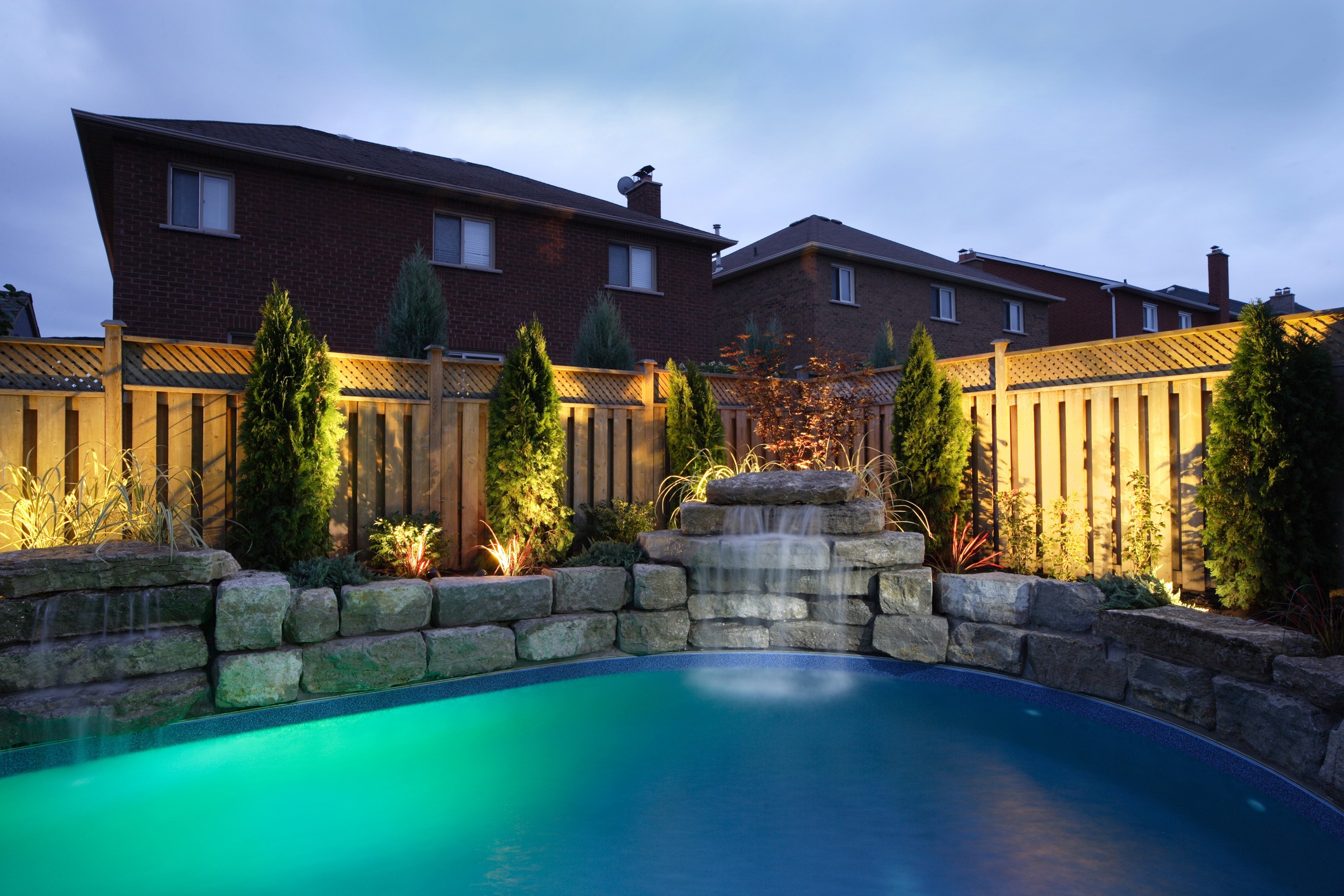 This photo shows an illuminated backyard with a swimming pool and waterfall at dusk, bordered by a wooden fence and landscaping, against a brick house background.