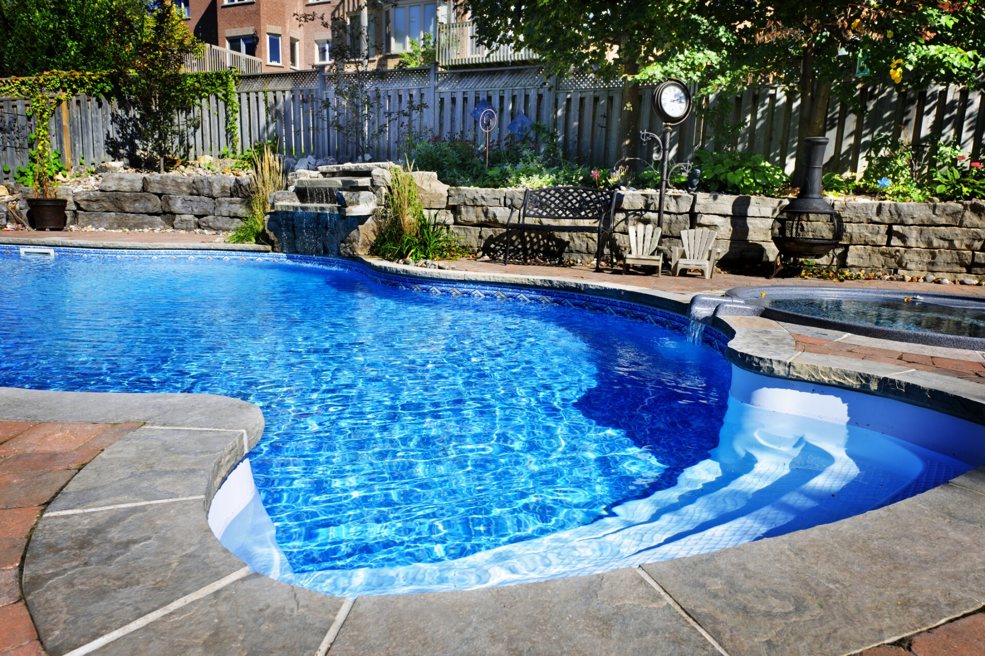 An inviting backyard with a curved in-ground swimming pool, stone patio, wooden fence, seating area, and a small waterfall on a sunny day.