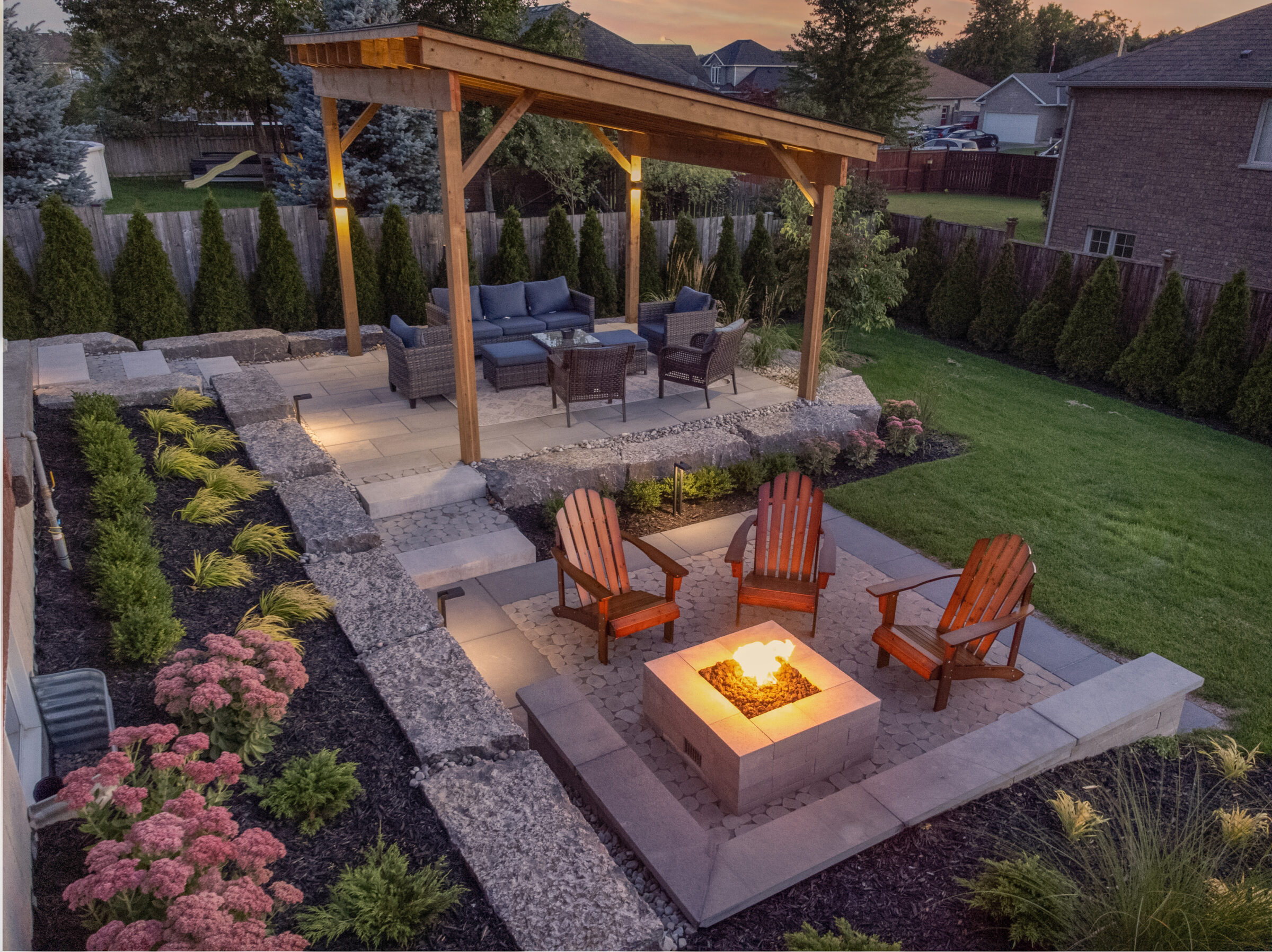 An elegantly landscaped backyard at dusk featuring a pergola, seating area, fire pit, stone pathways, manicured grass, and ornamental plants.