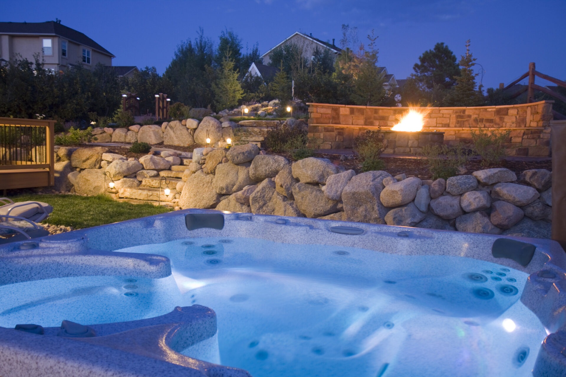 A luxurious hot tub sits in a beautifully landscaped backyard at dusk, with atmospheric lighting and a rock fire pit glowing in the background.