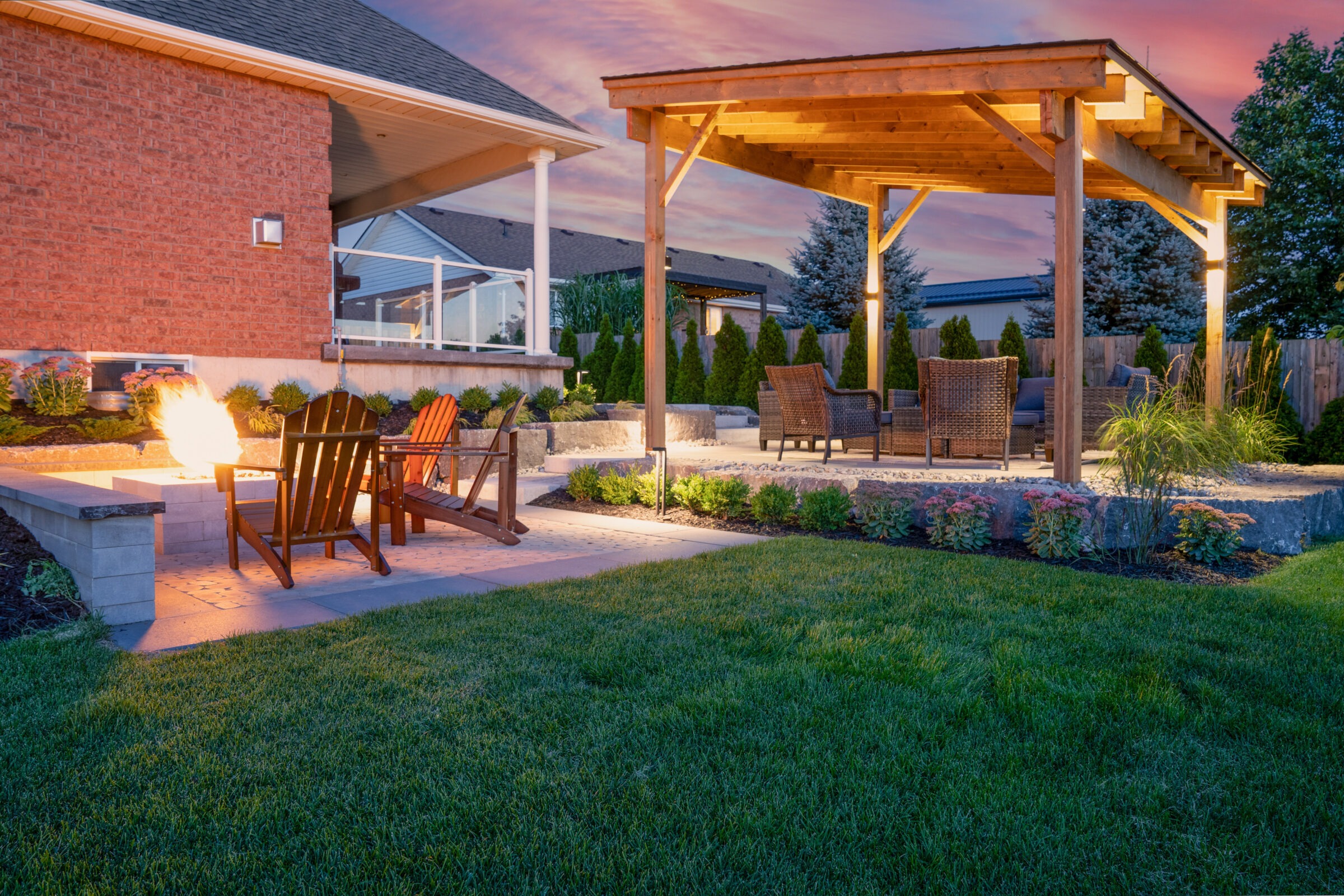 This image showcases a beautifully landscaped backyard during dusk with a fire pit, Adirondack chairs, a covered patio, well-maintained lawn, and garden lighting.