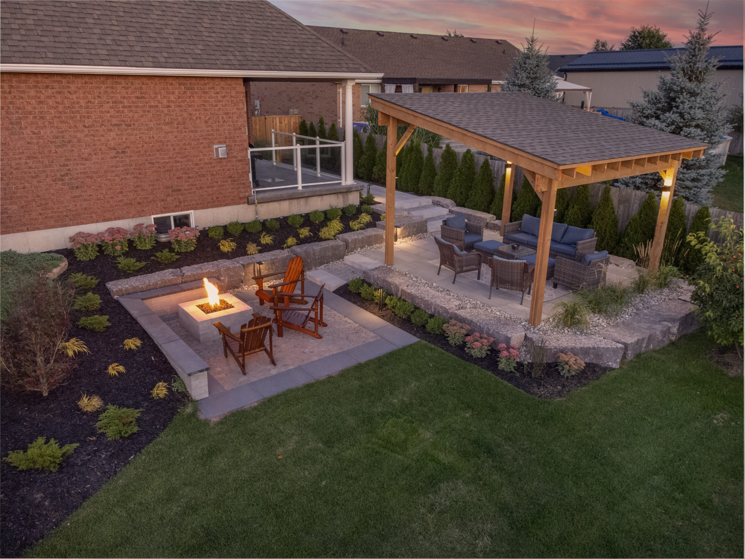 An outdoor living space with a fire pit, wooden chairs, a covered seating area, landscaping, and a brick house during twilight.