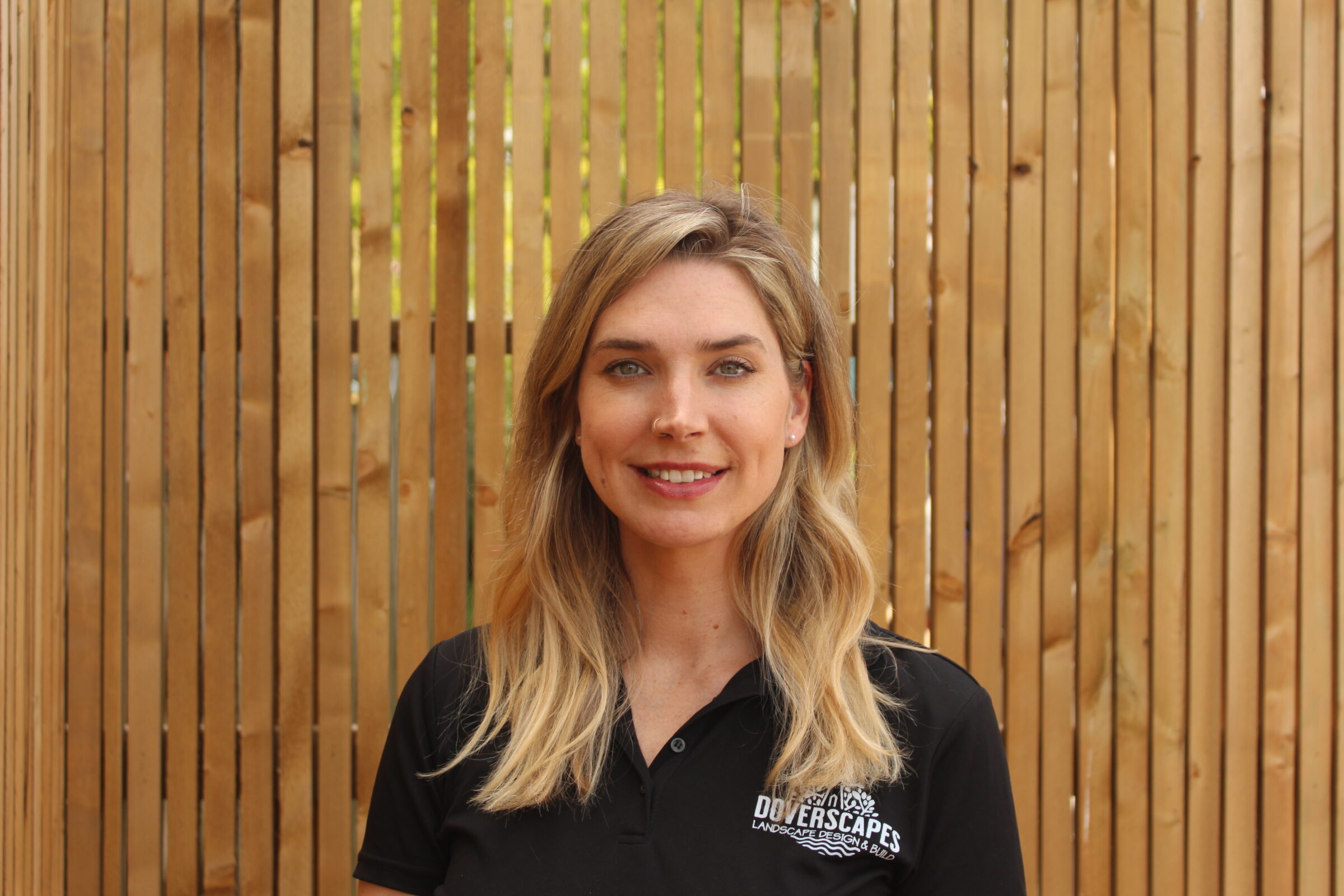 A person with blond hair and a black polo shirt is smiling, standing in front of a wooden slat background in a well-lit setting.