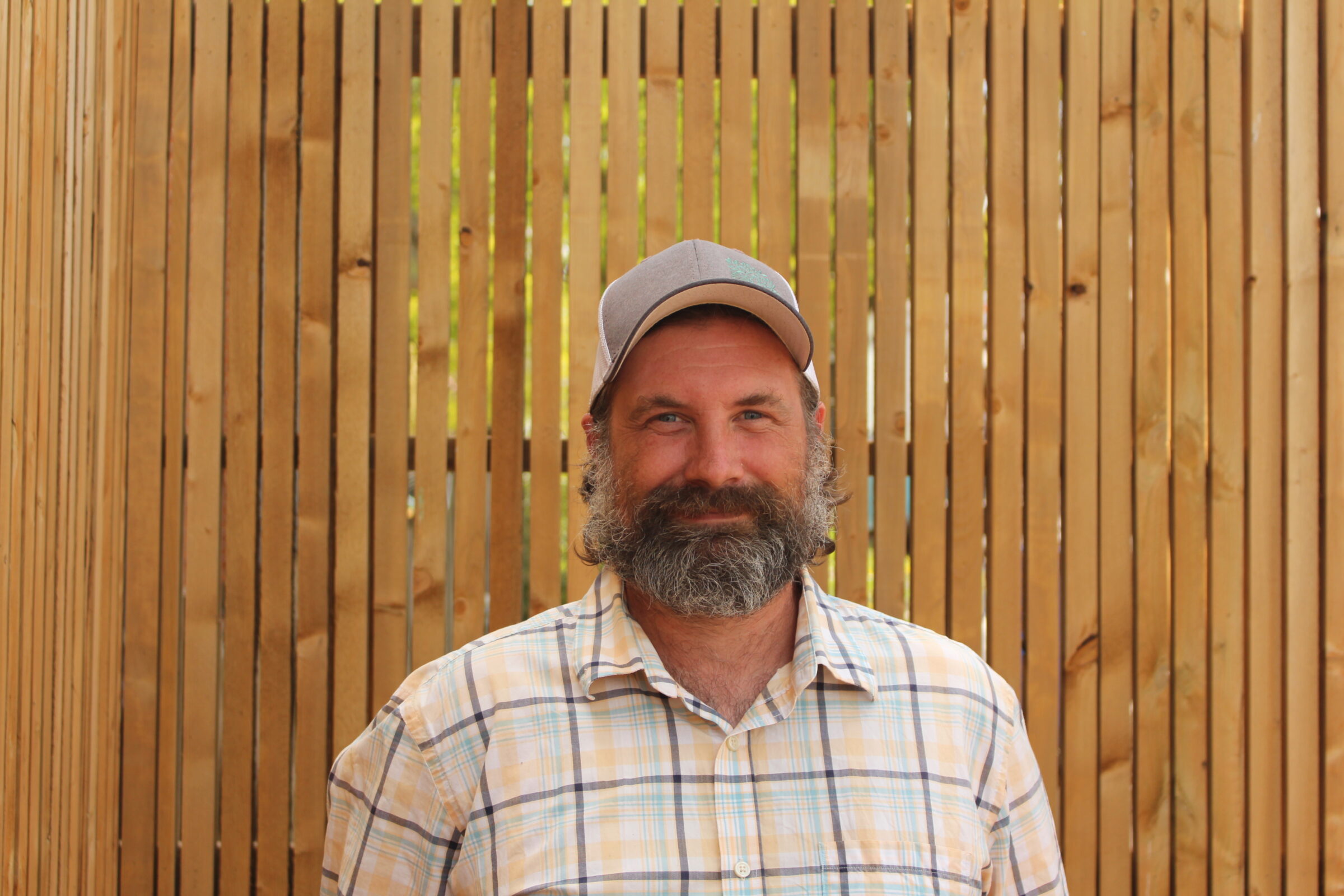 A person with a beard and cap smiles in front of a wooden slat background. They're wearing a plaid shirt and appear content. The setting seems casual.