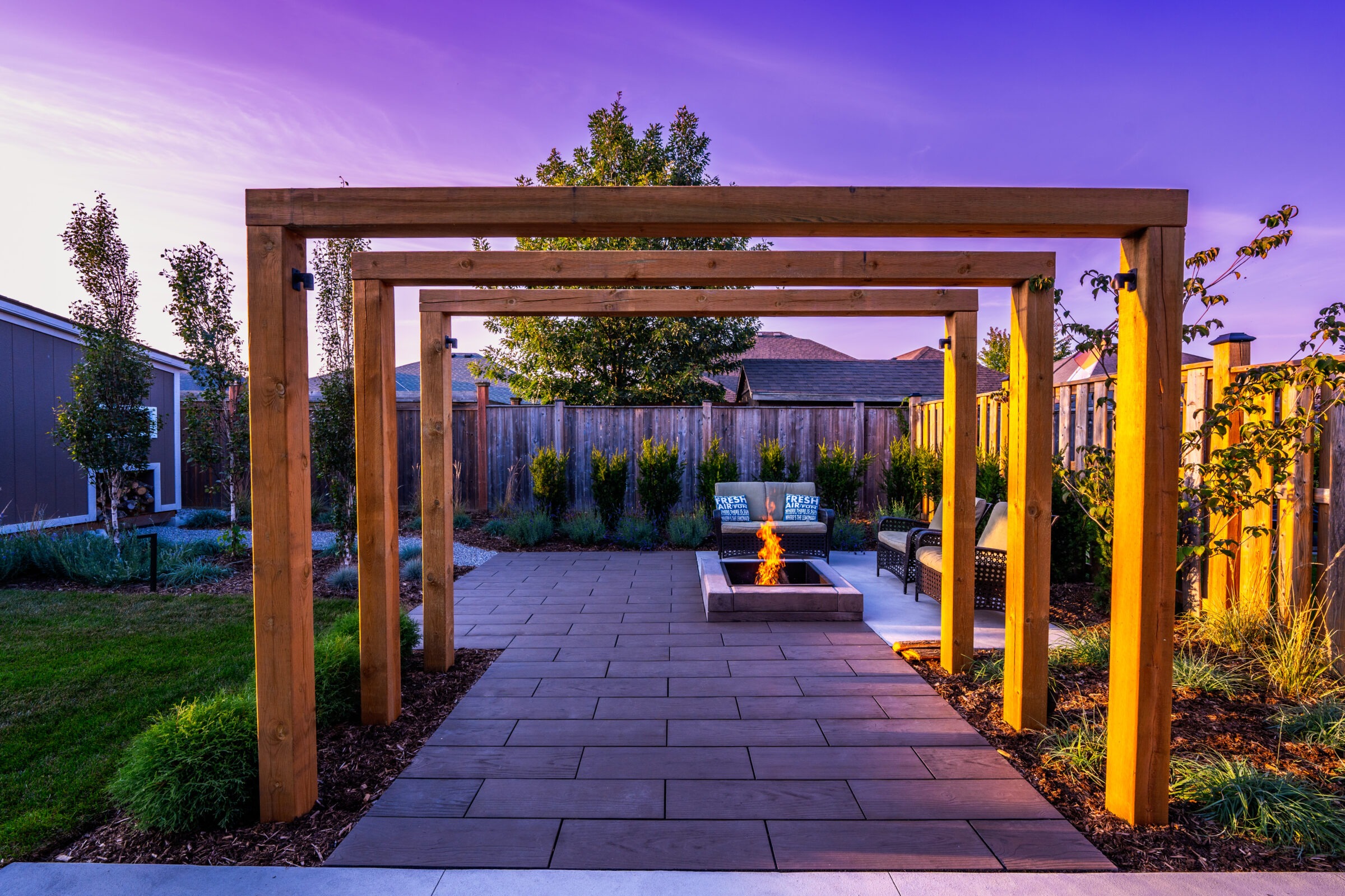 A serene backyard at dusk featuring a wooden pergola, a lit fire pit, arranged seating, well-maintained grass, and a pathway under a colorful sky.