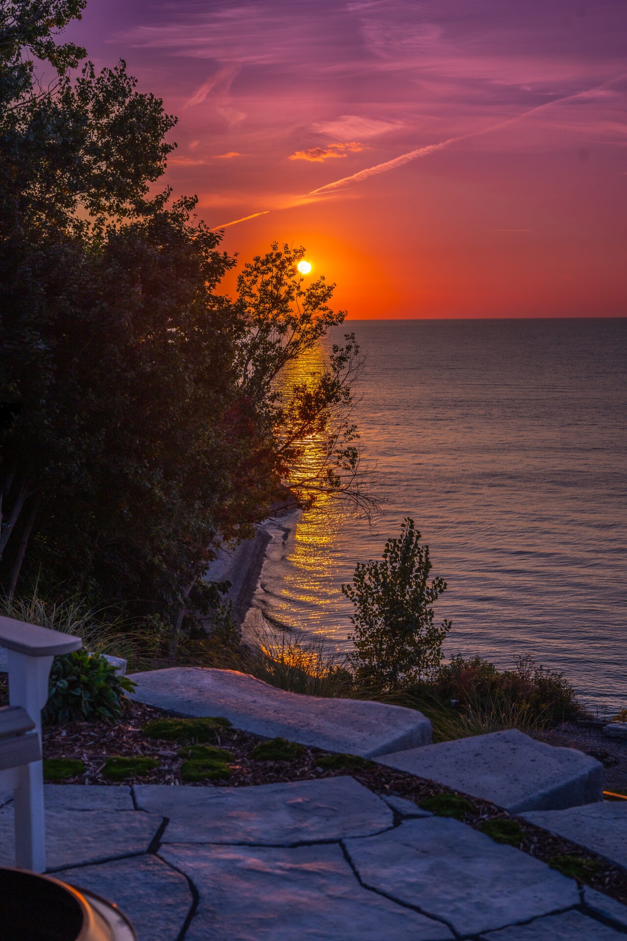 A tranquil sunset view over a calm sea with the sun casting golden reflections on the water, stone steps lead towards a tree-lined shore.