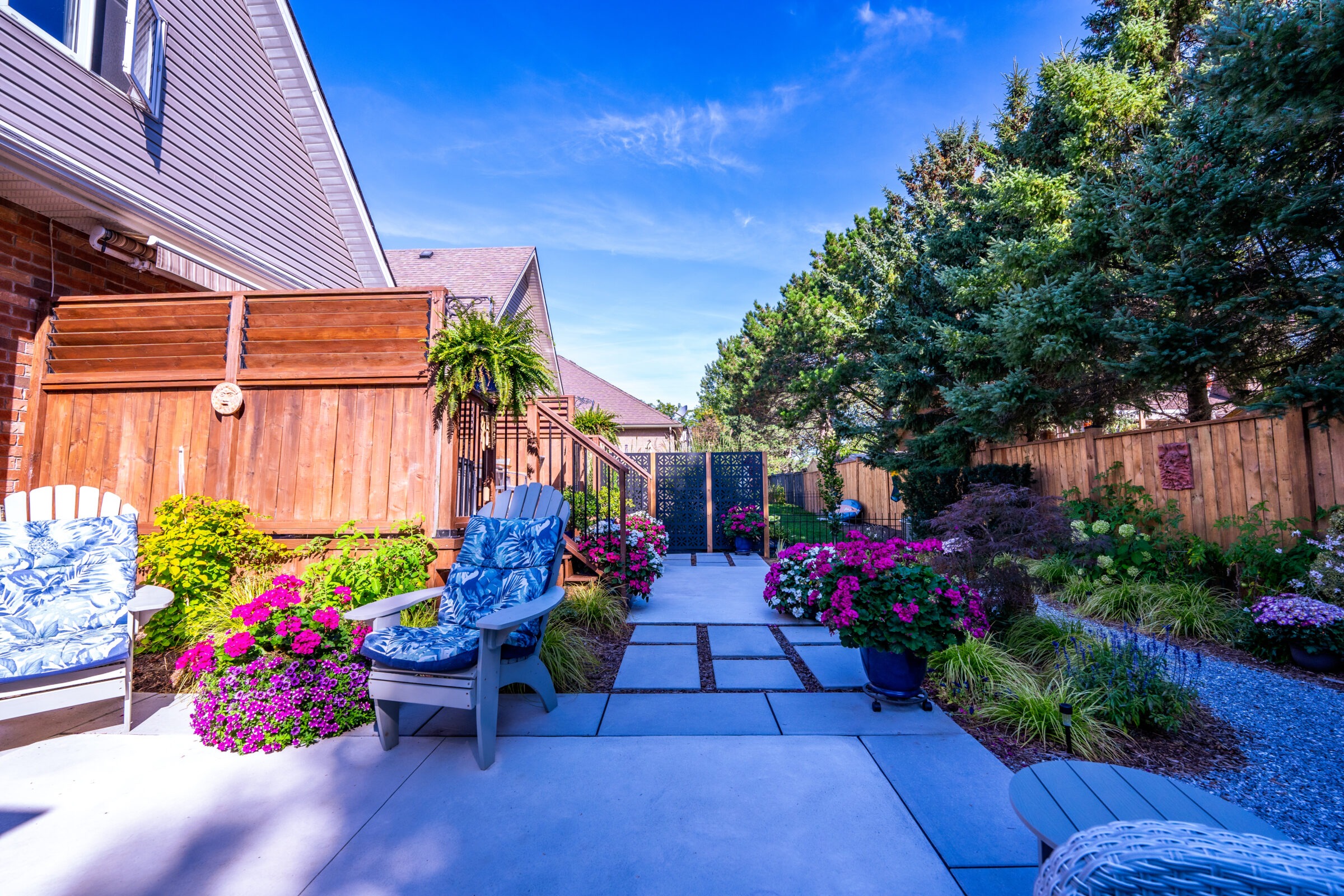 A well-manicured garden path lined with vibrant flowers leads to a decorative gate, flanked by lush greenery and comfortable seating under a clear blue sky.