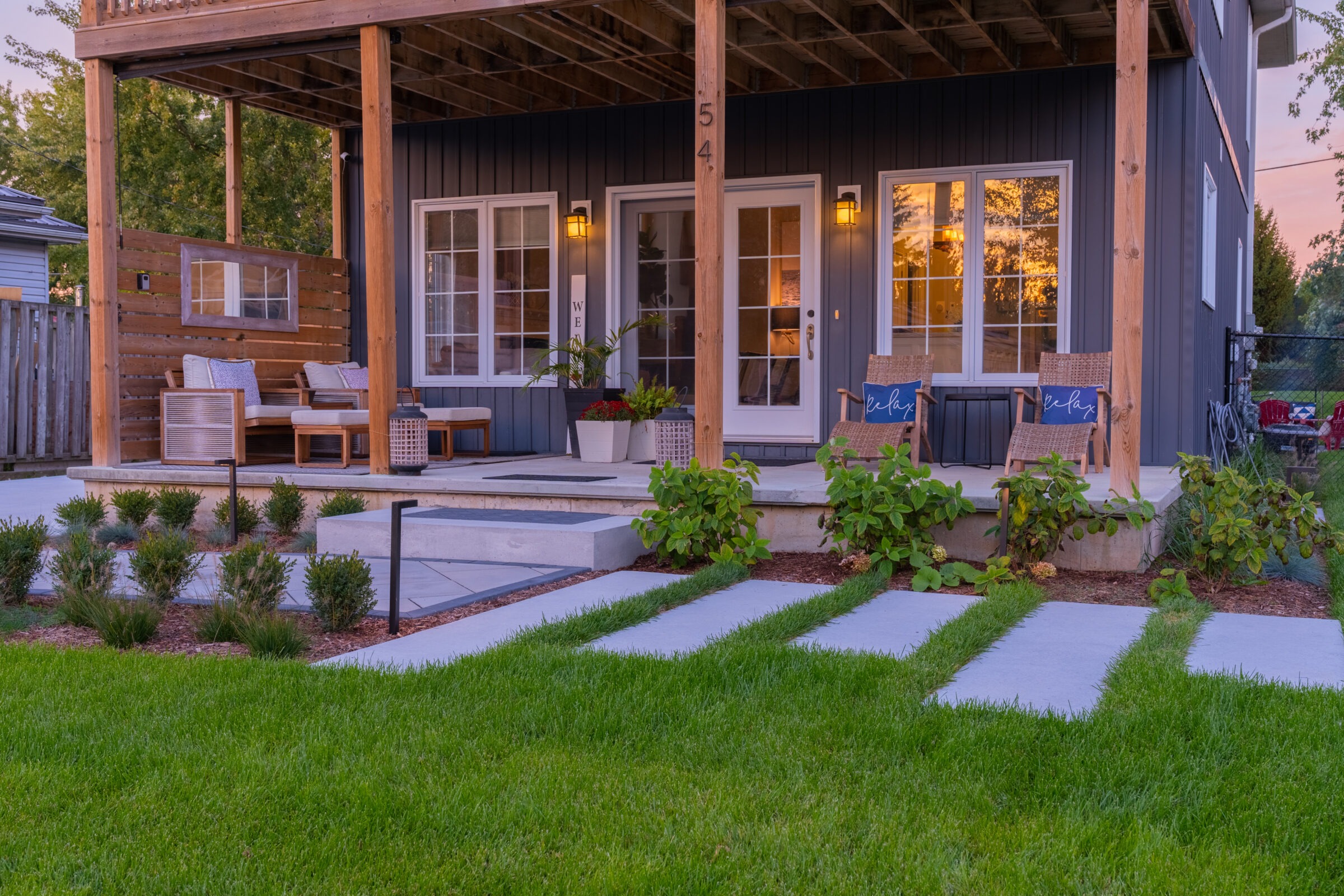A cozy porch with seating and plants outside a blue house with large windows reflecting a sunset, leading to a well-manicured lawn with stepping stones.