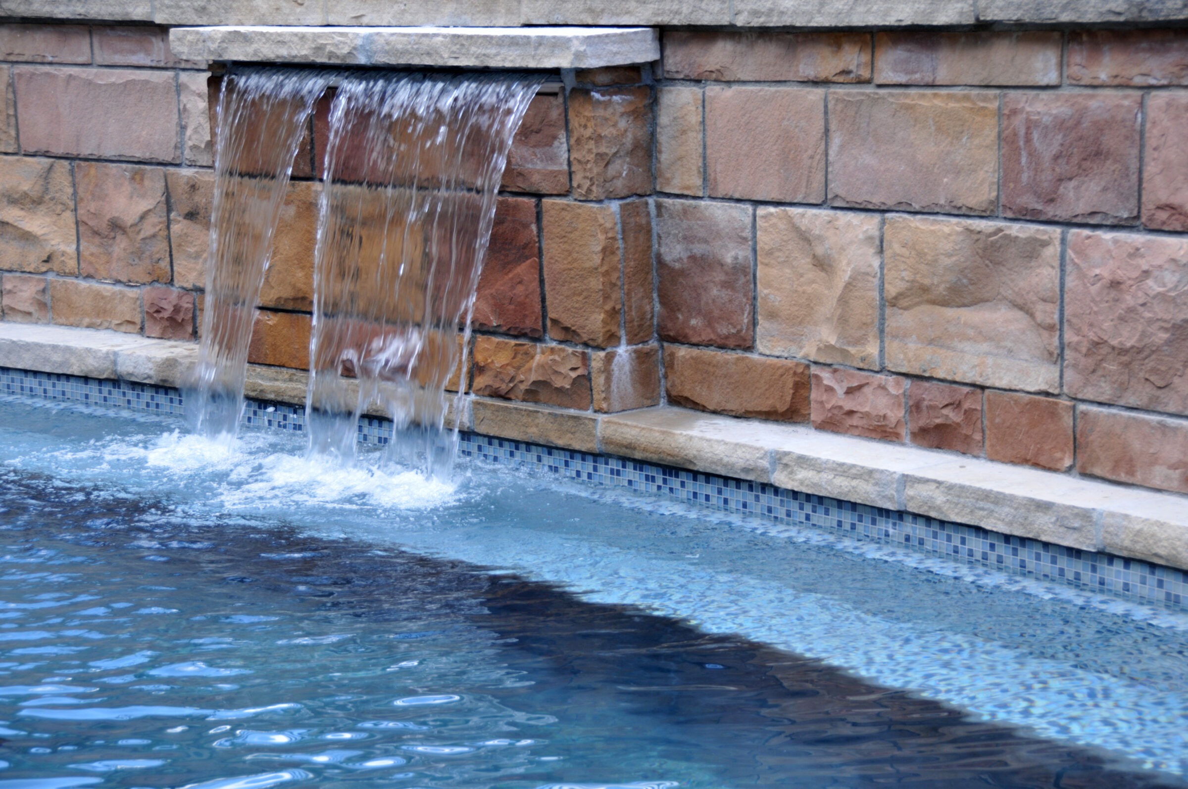 The image shows a water feature with cascading water from a stone wall into a pool with blue mosaic tiles. The water looks serene and inviting.